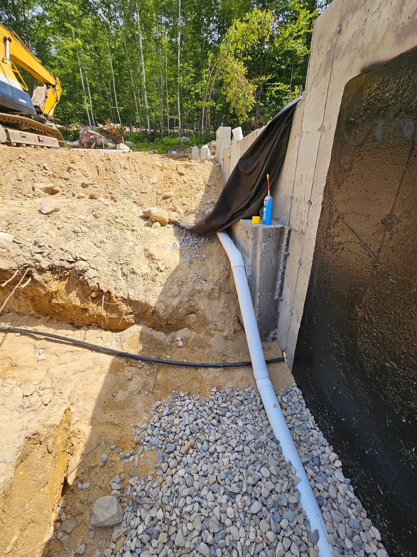 Construction site: trench with drainpipe against a concrete wall. Black tarp and excavator visible.