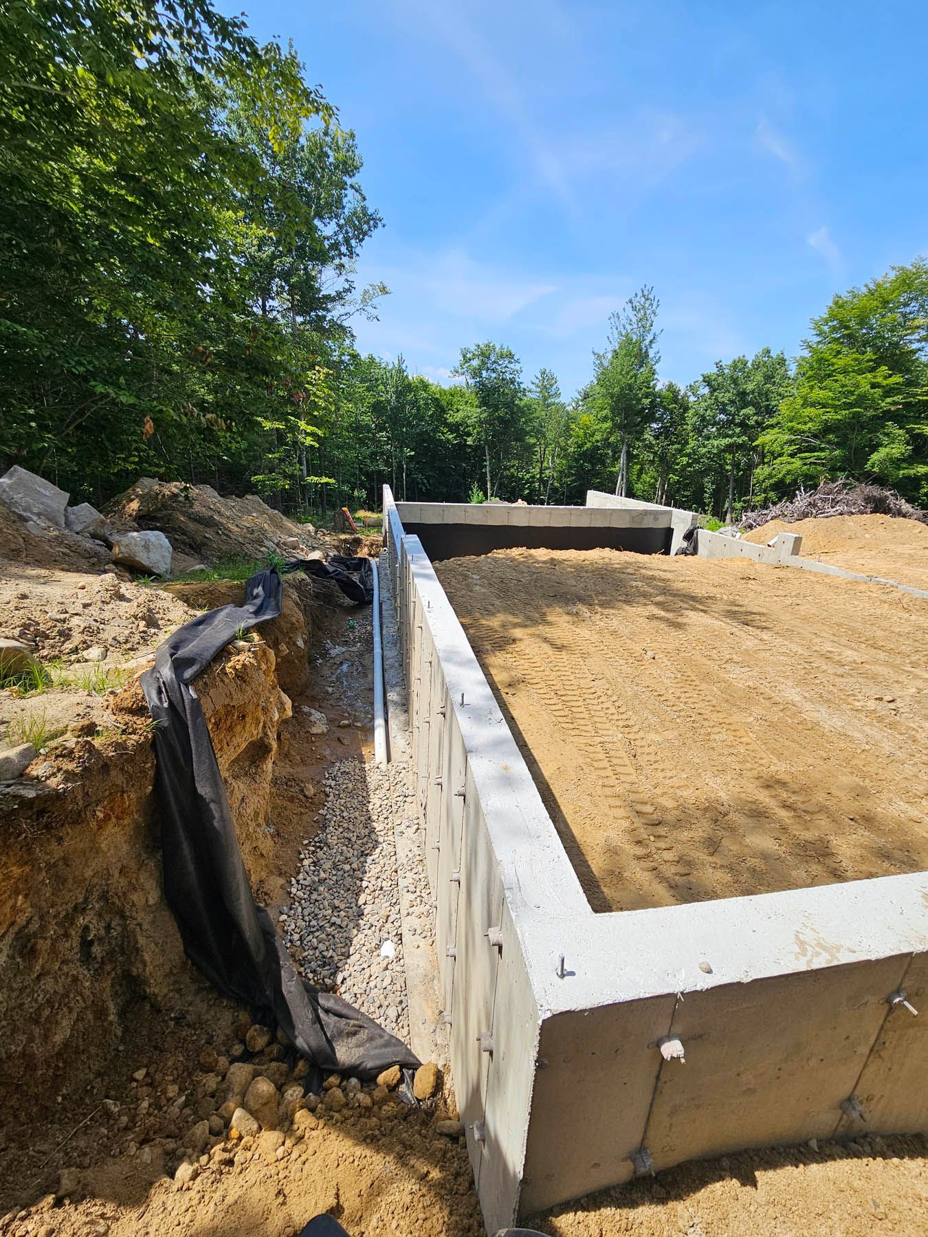 Foundation of a building under construction; concrete walls, dirt fill, surrounding trees, and blue sky.