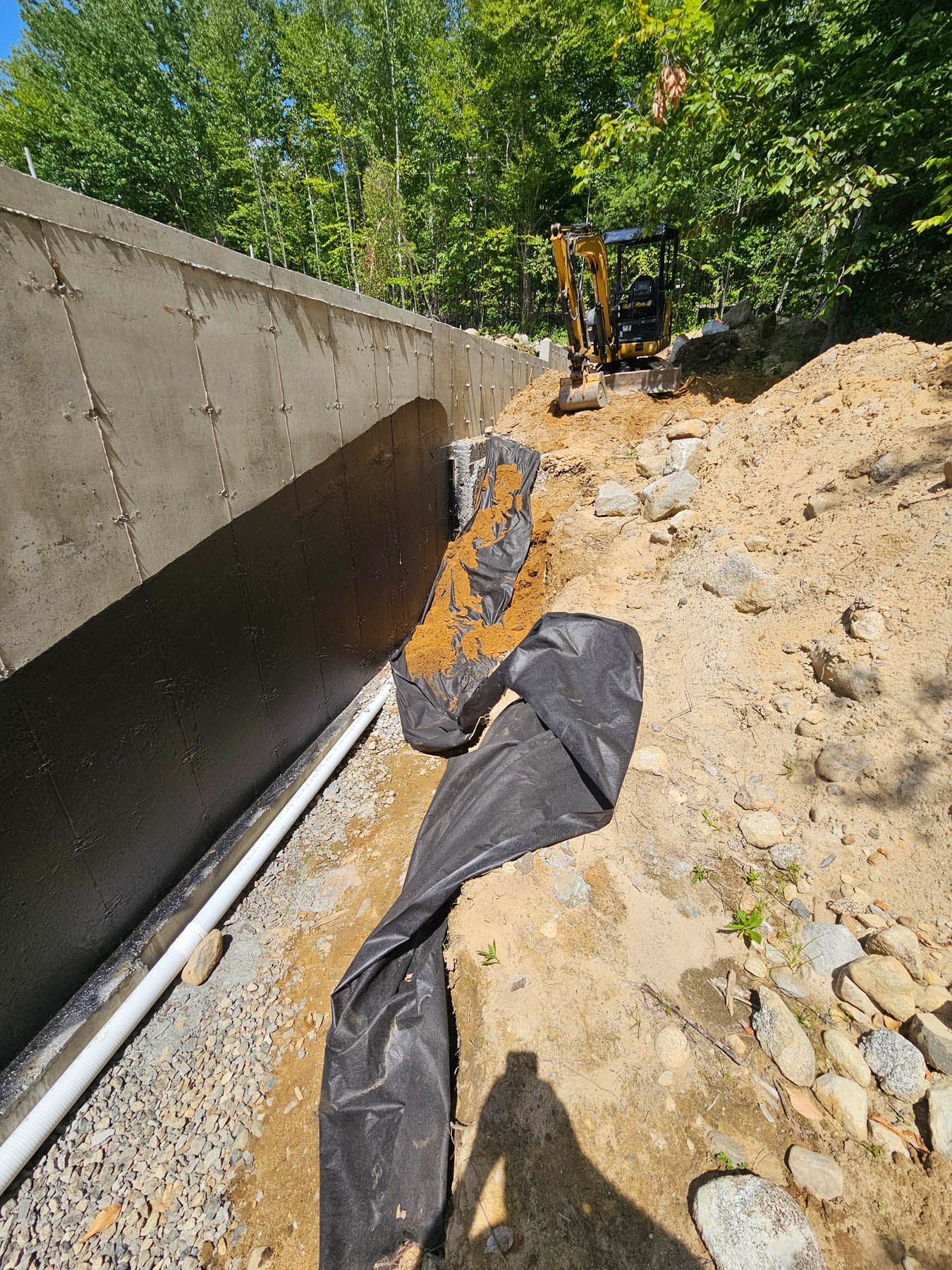 Construction site with a foundation wall, drainage pipe, and excavator. A black fabric covers the soil.