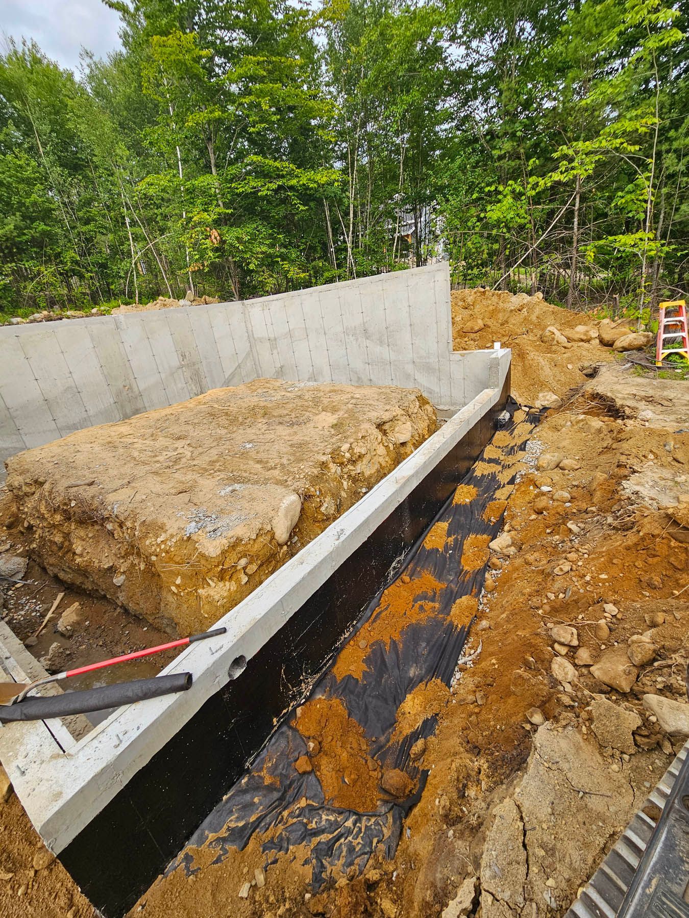 Construction site: foundation wall with excavated soil, black lining, and a ladder in the background.