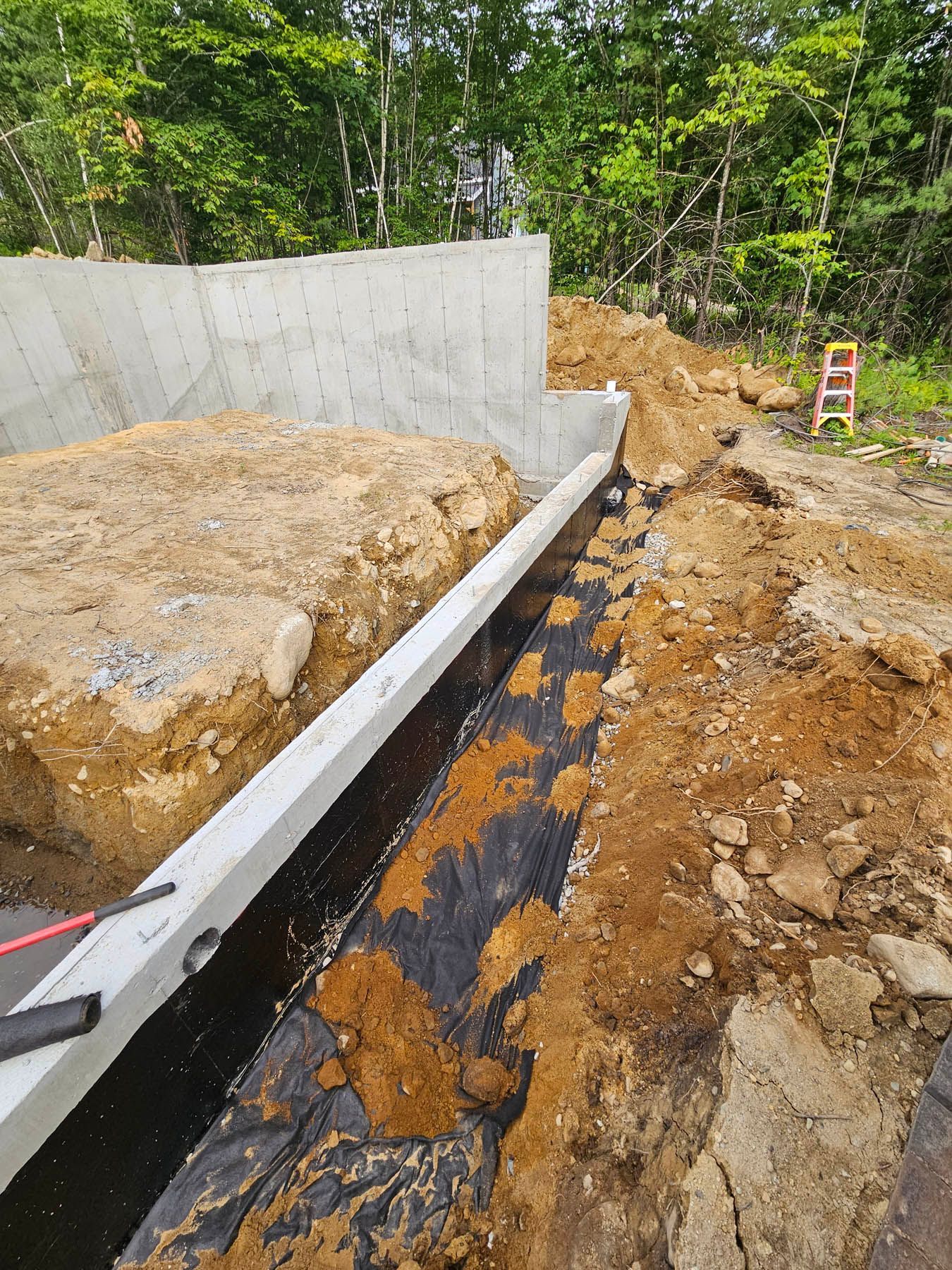 Construction site with concrete foundation, trench, and black fabric. Soil, trees, and orange sign are visible.