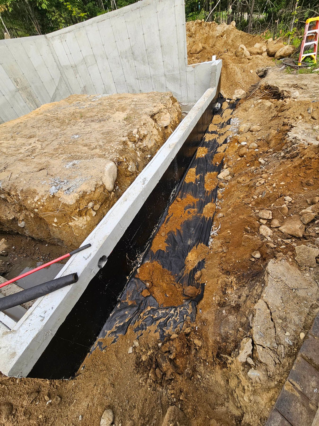 Construction site with a trench dug along a concrete foundation wall, lined with black fabric.