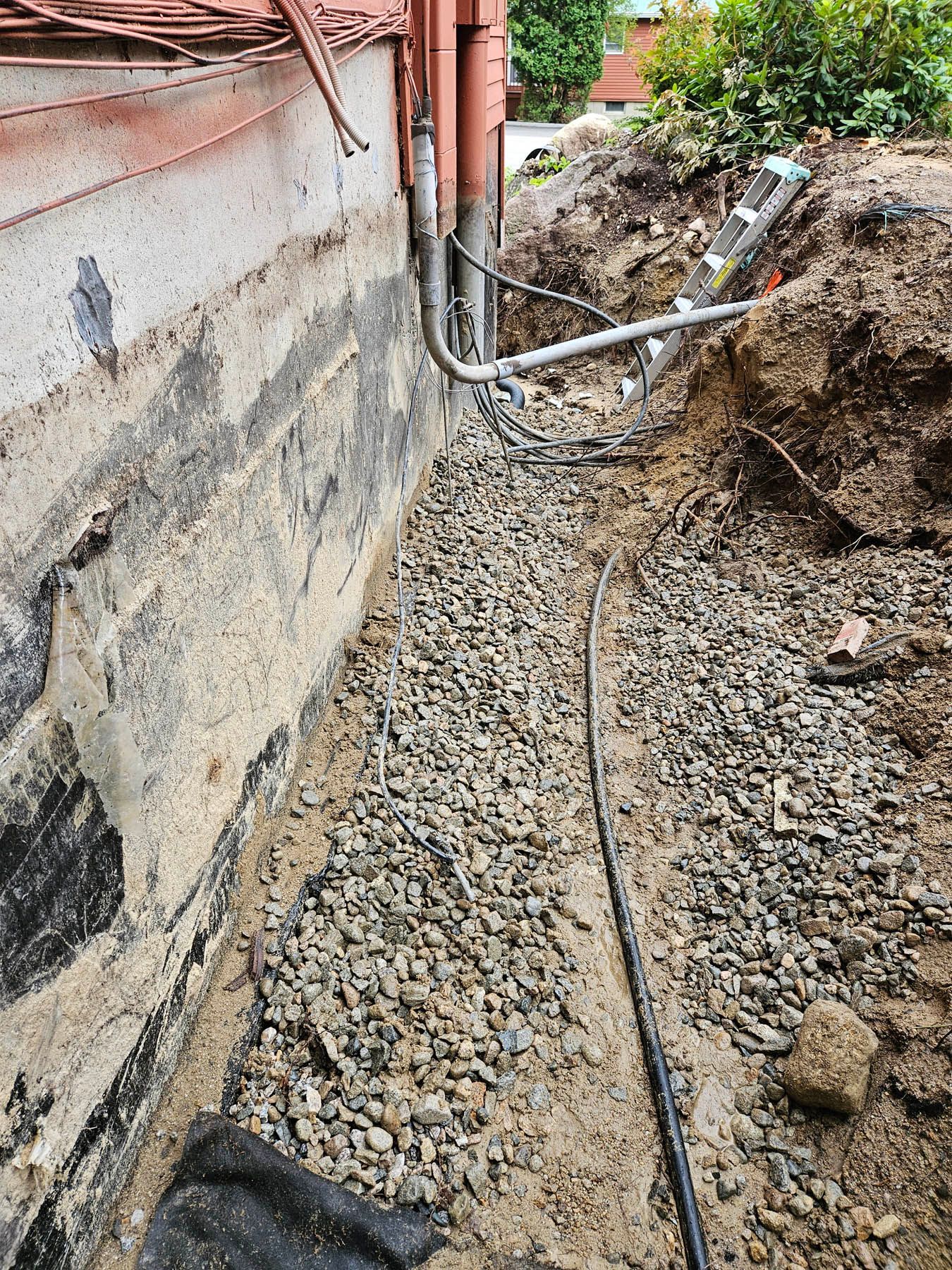 Trench dug alongside a building's foundation, filled with gravel, electrical conduit and pipe visible.