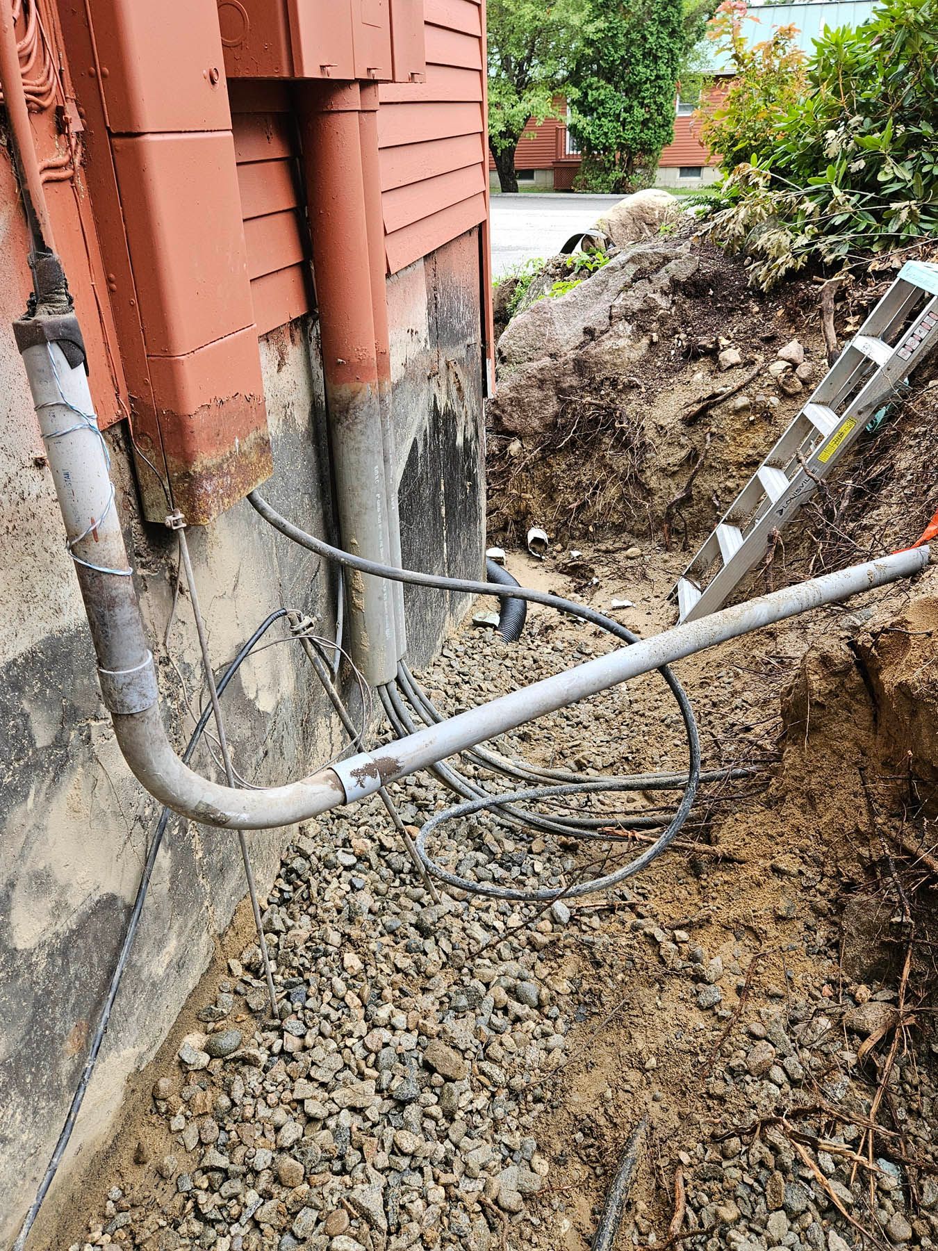 Foundation work with exposed pipes next to a red-painted building. A ladder leans against an excavated dirt pile.