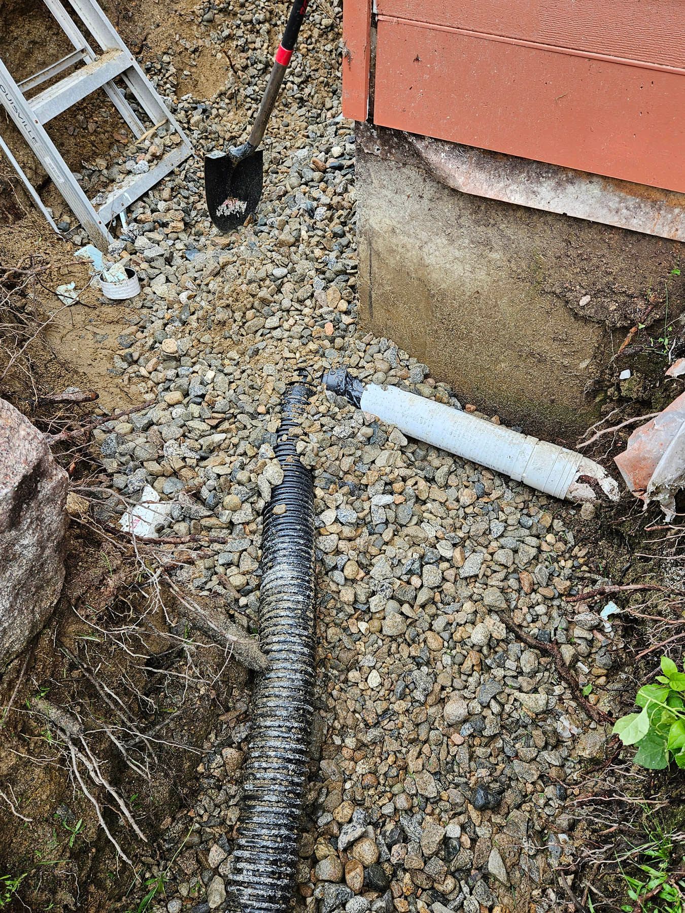 Drainage system installation: black corrugated pipe in a gravel trench near a building's foundation.