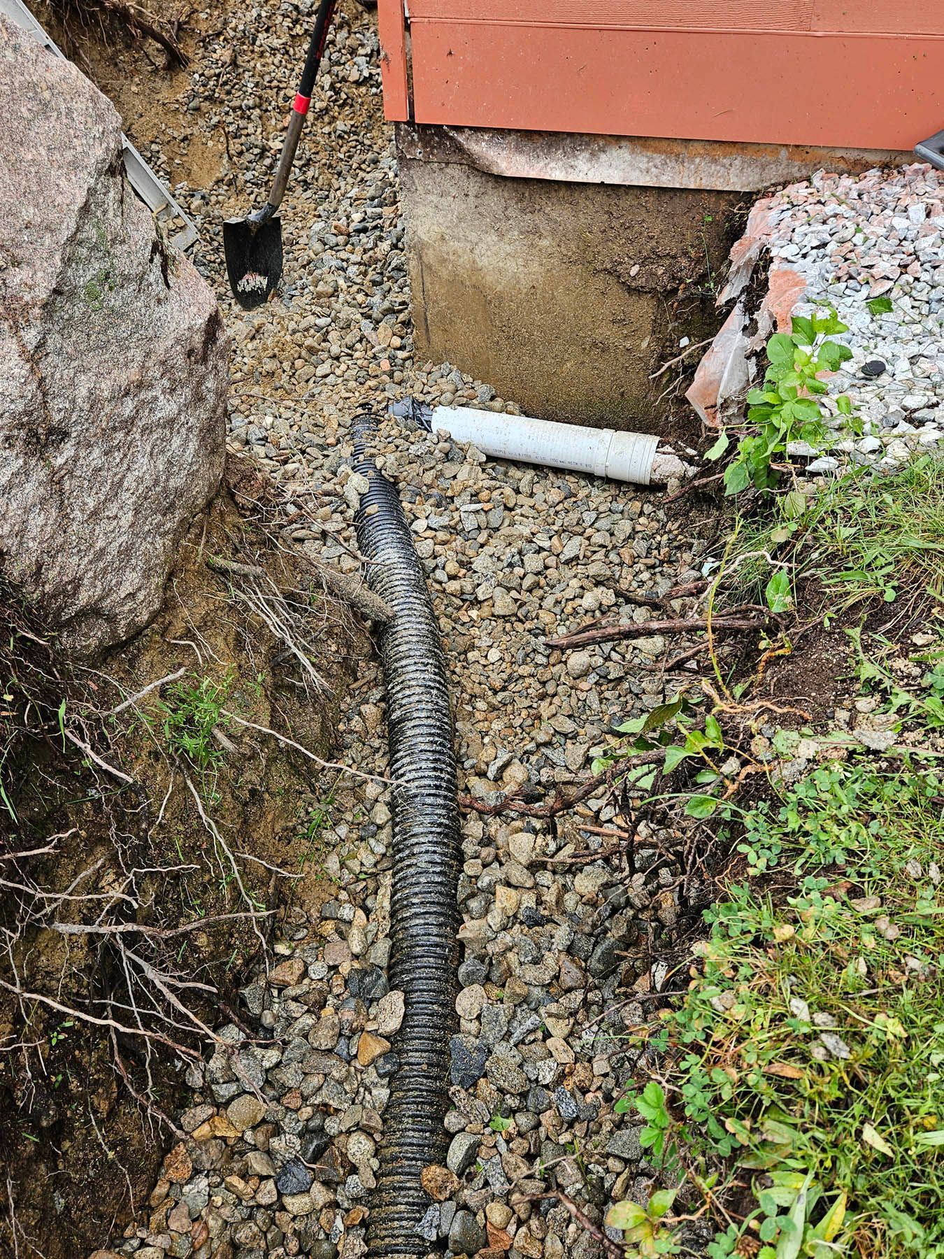 Drainage system installation with perforated pipe in a gravel trench near a building foundation and rocks.