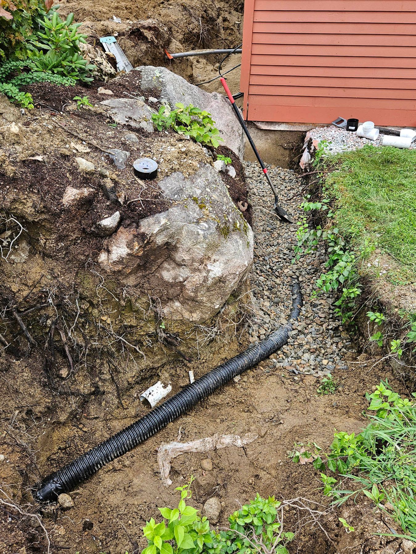 Construction site with drainage pipe, gravel, large rock, and red building wall.