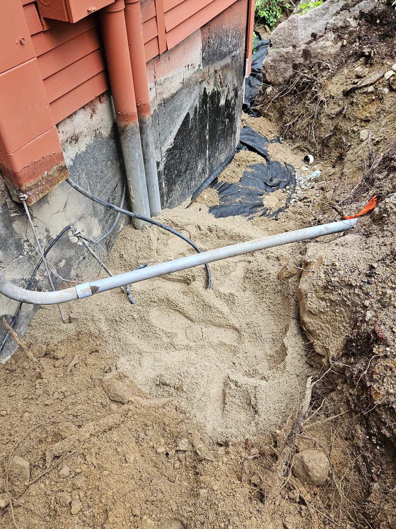 Trench dug alongside a red house's foundation with pipes and electrical conduit visible.