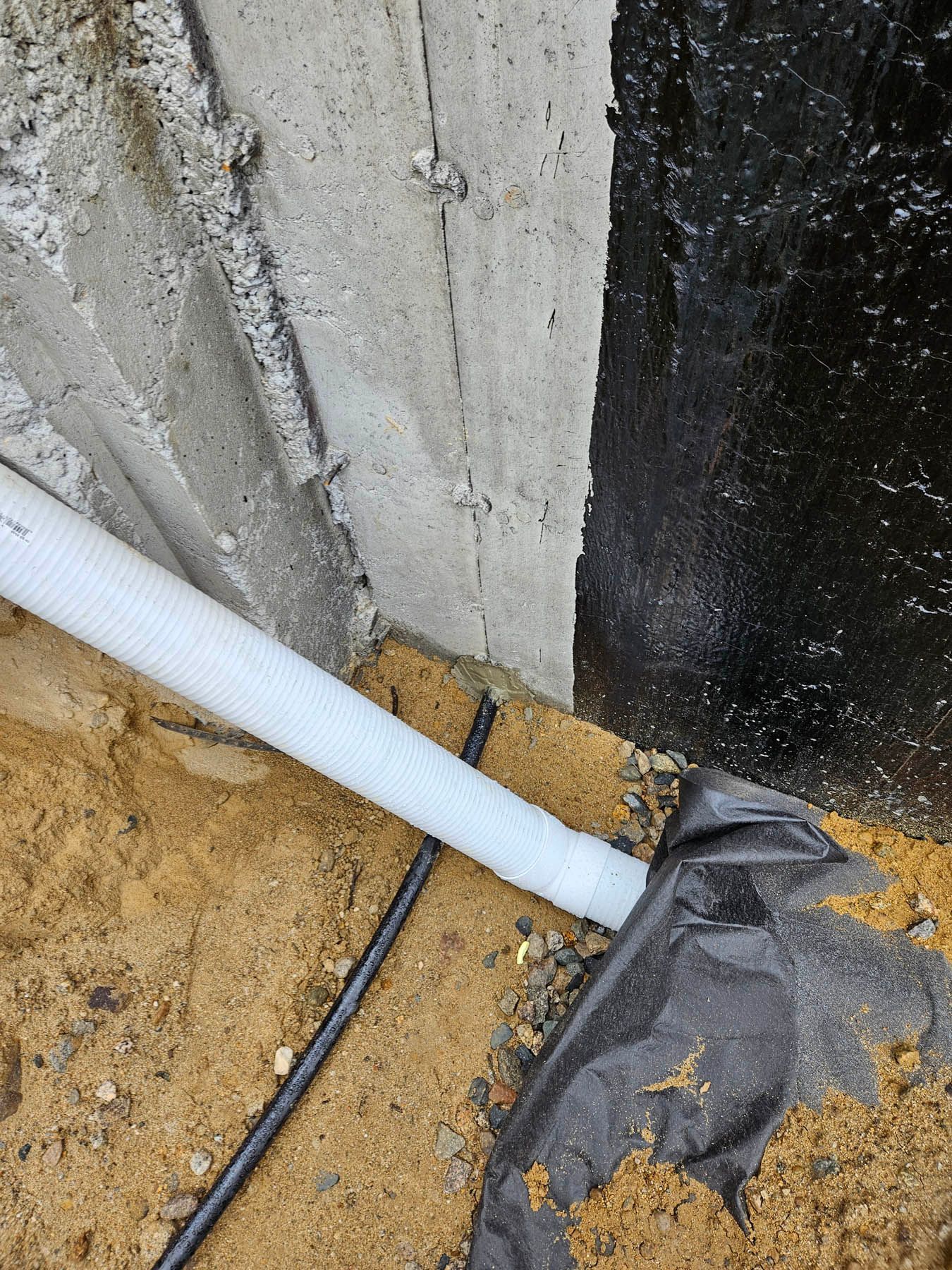 Foundation drainage system with white pipe, black waterproofing, and soil.