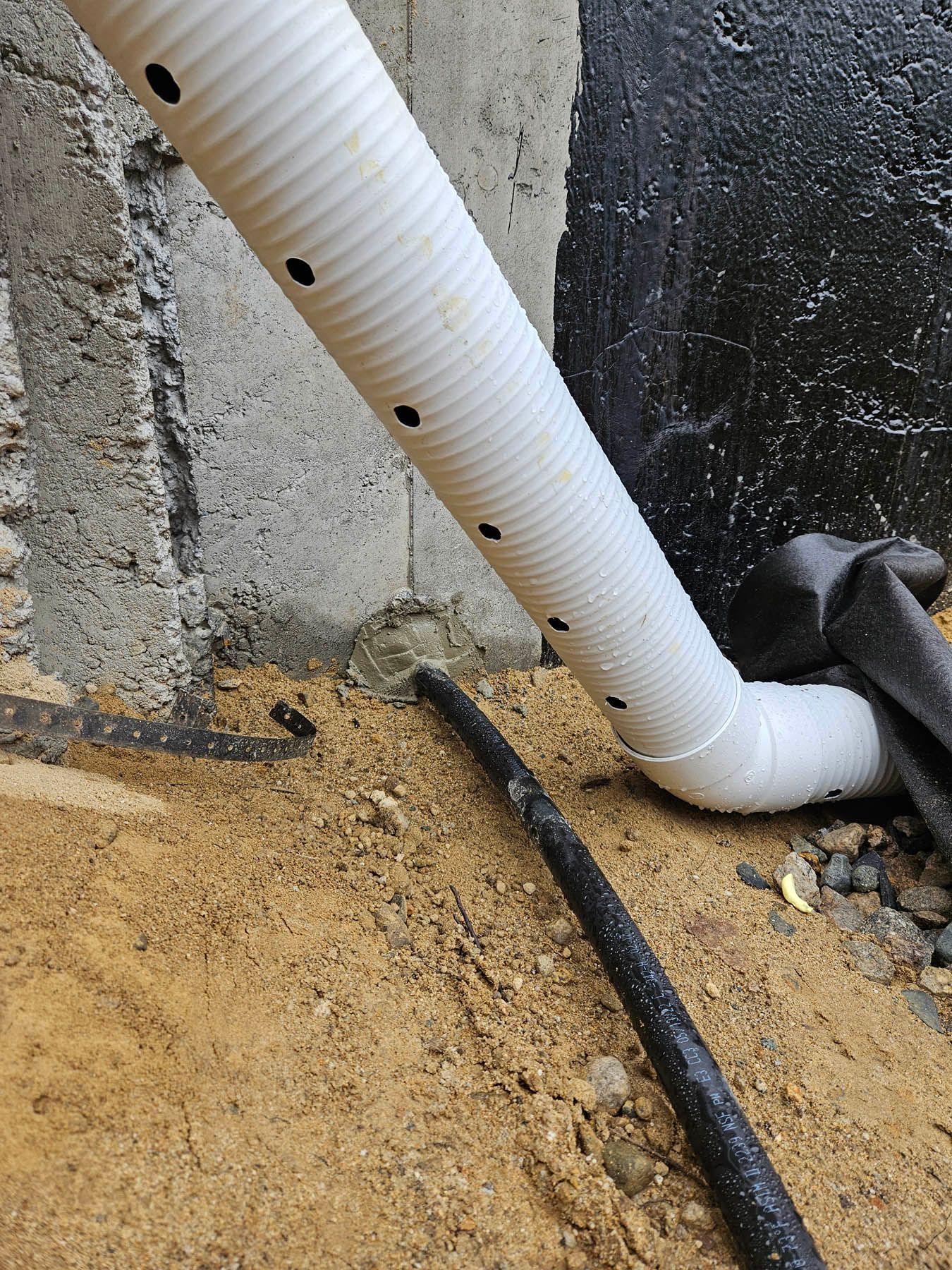 White and black drain pipes against a concrete wall. The wall is partially covered in a black sealant and resting on dirt.