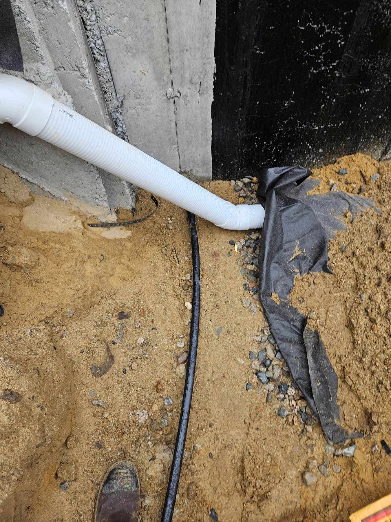 White drain pipe connected to a black fabric-covered drainage system in a sandy area, next to a concrete wall.