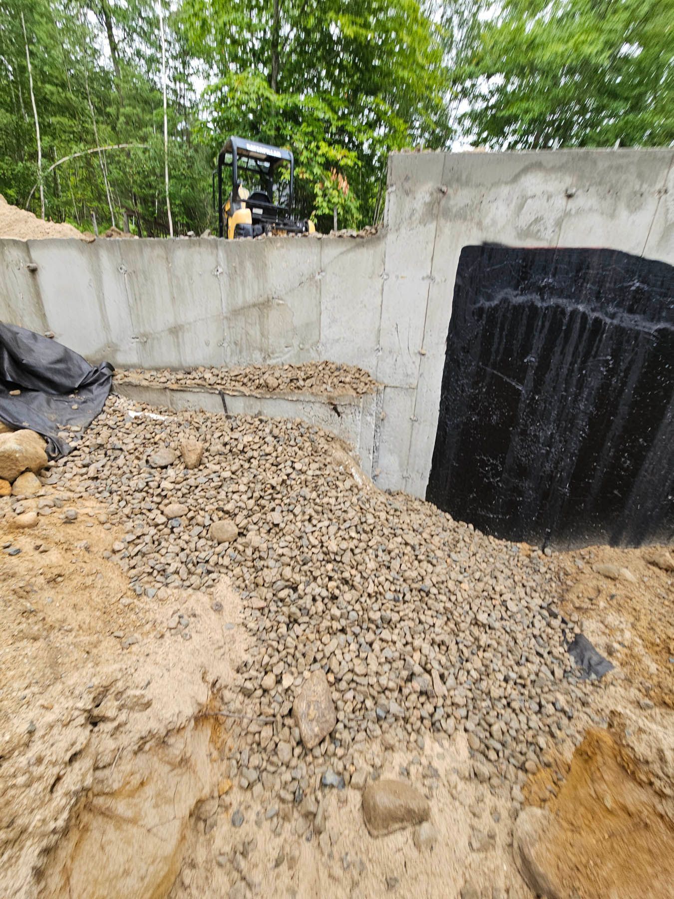 Construction site with concrete wall, gravel, and black waterproofing membrane; a mini excavator is in the background.