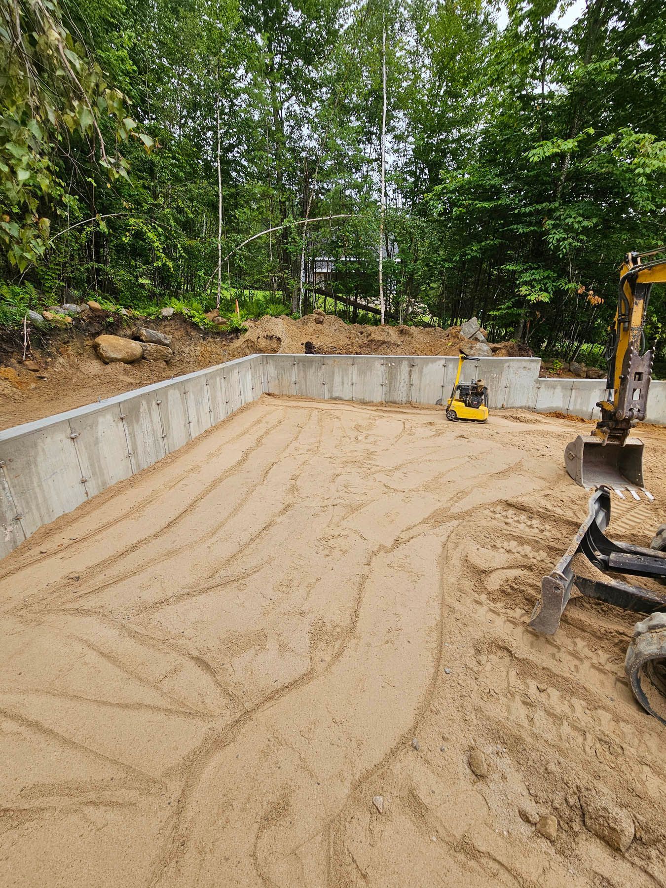 Construction site with concrete foundation and earth. Mini excavator on the right. Forest backdrop.