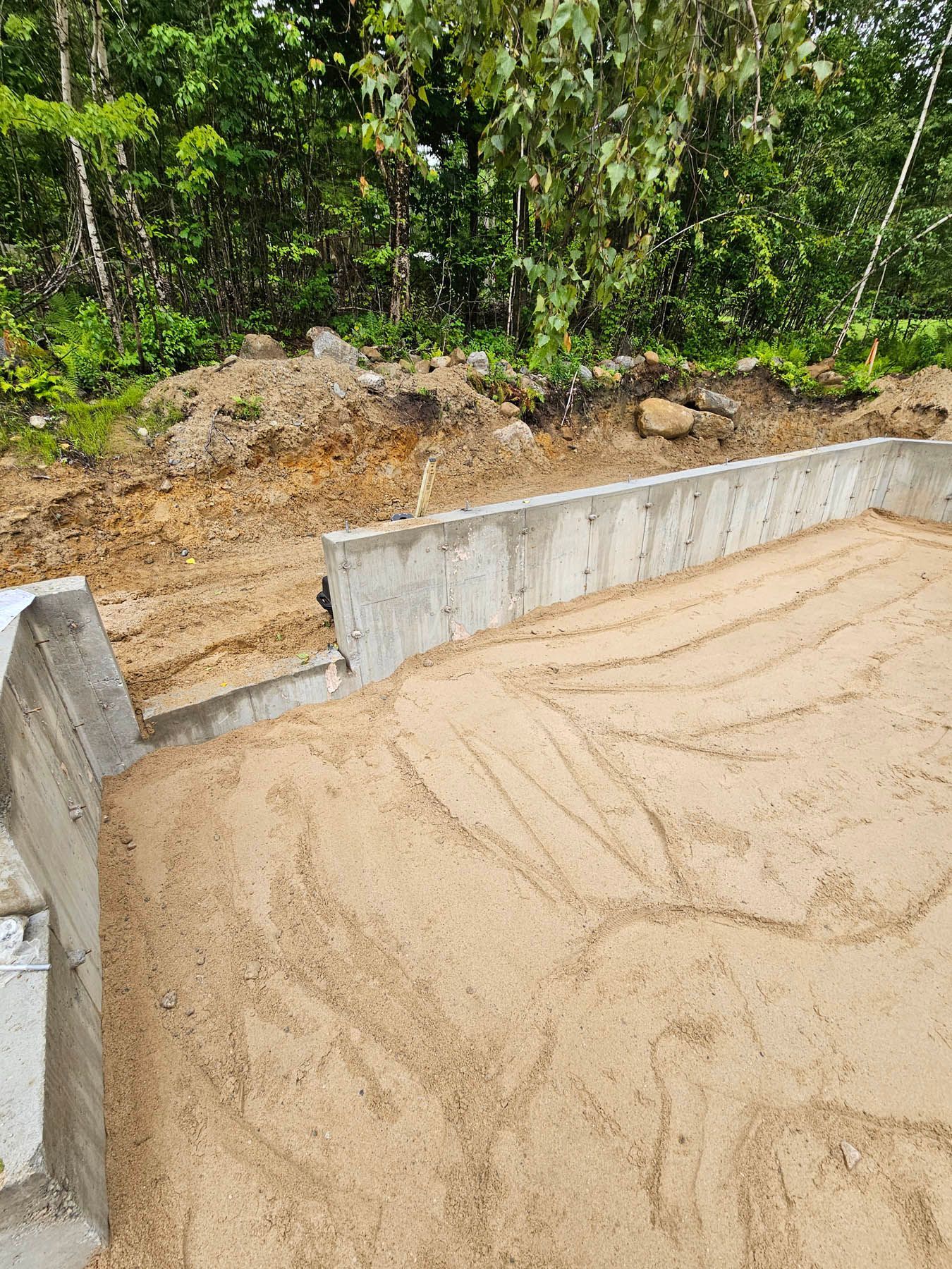 Construction site with concrete walls and sand; trees in the background.