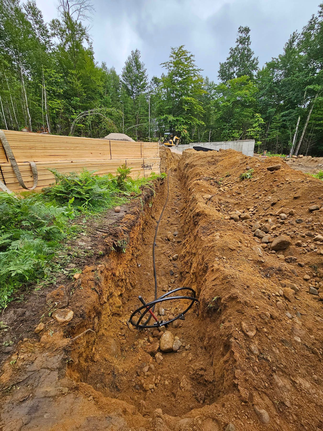 Trench dug in brown earth with black cable. Green trees and construction site in the background.