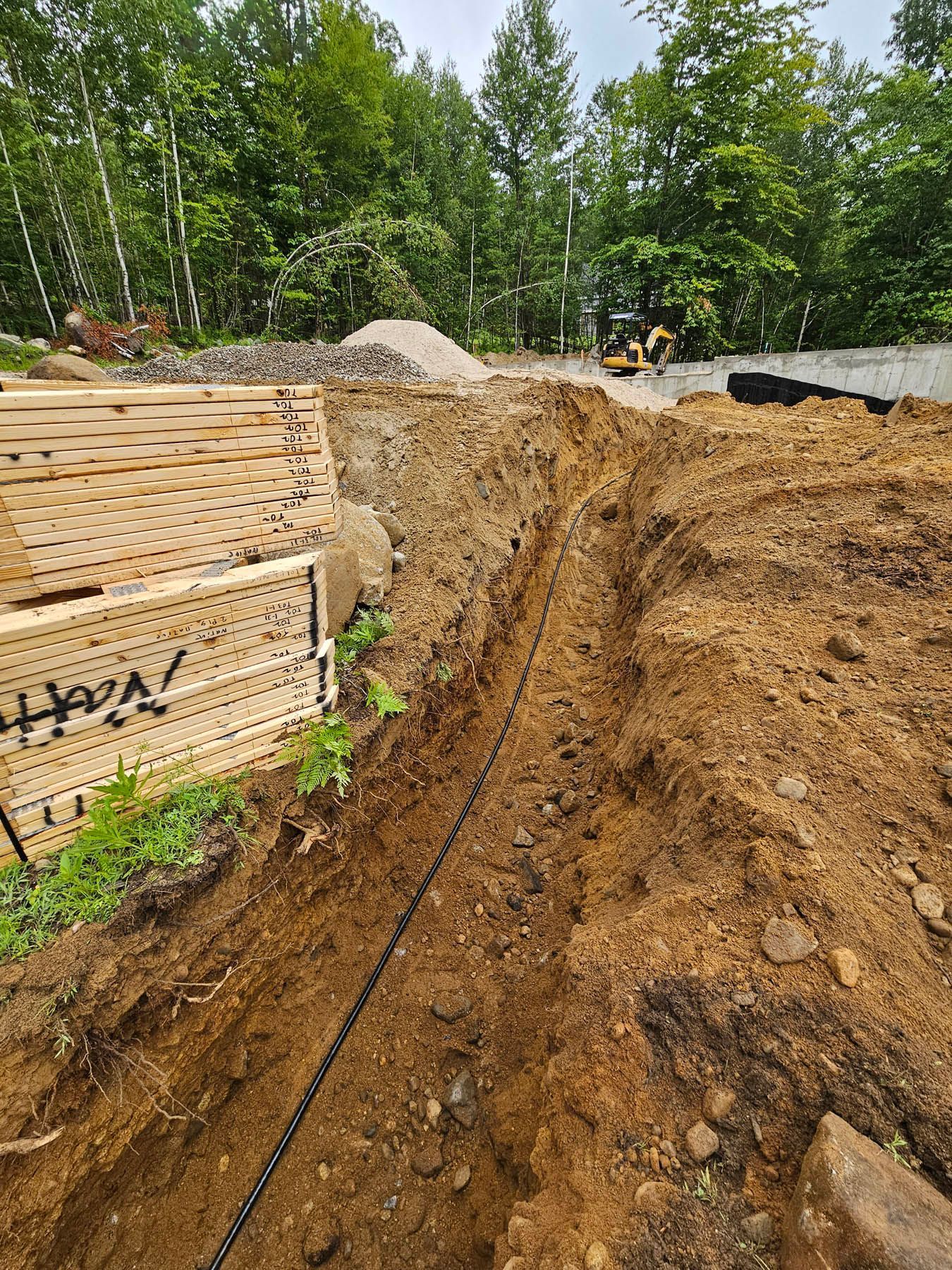 Trench dug in dirt with black cable, lumber, gravel, and small excavator in the background.