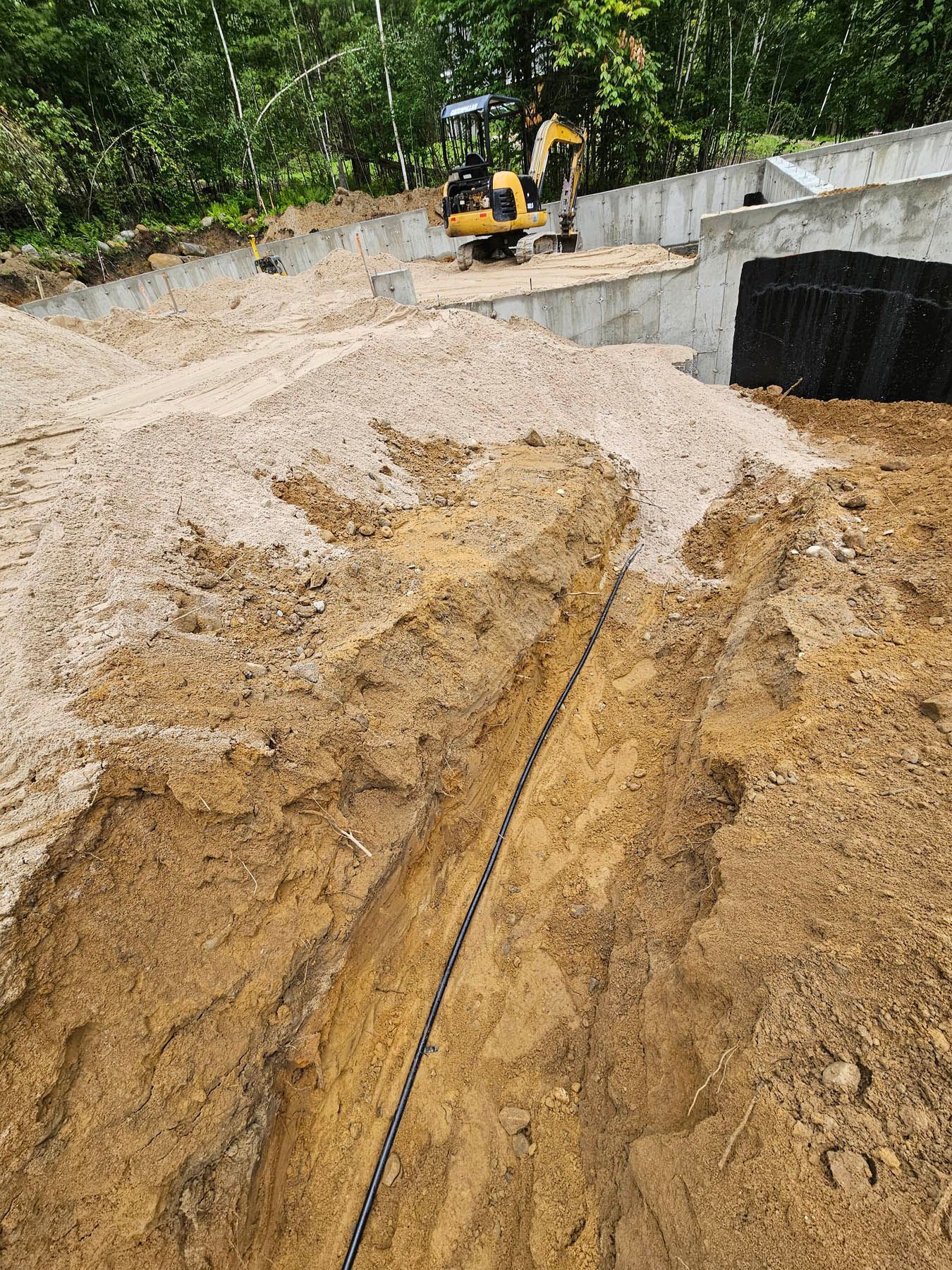 Construction site with excavated trench, backhoe, soil, and concrete retaining walls.