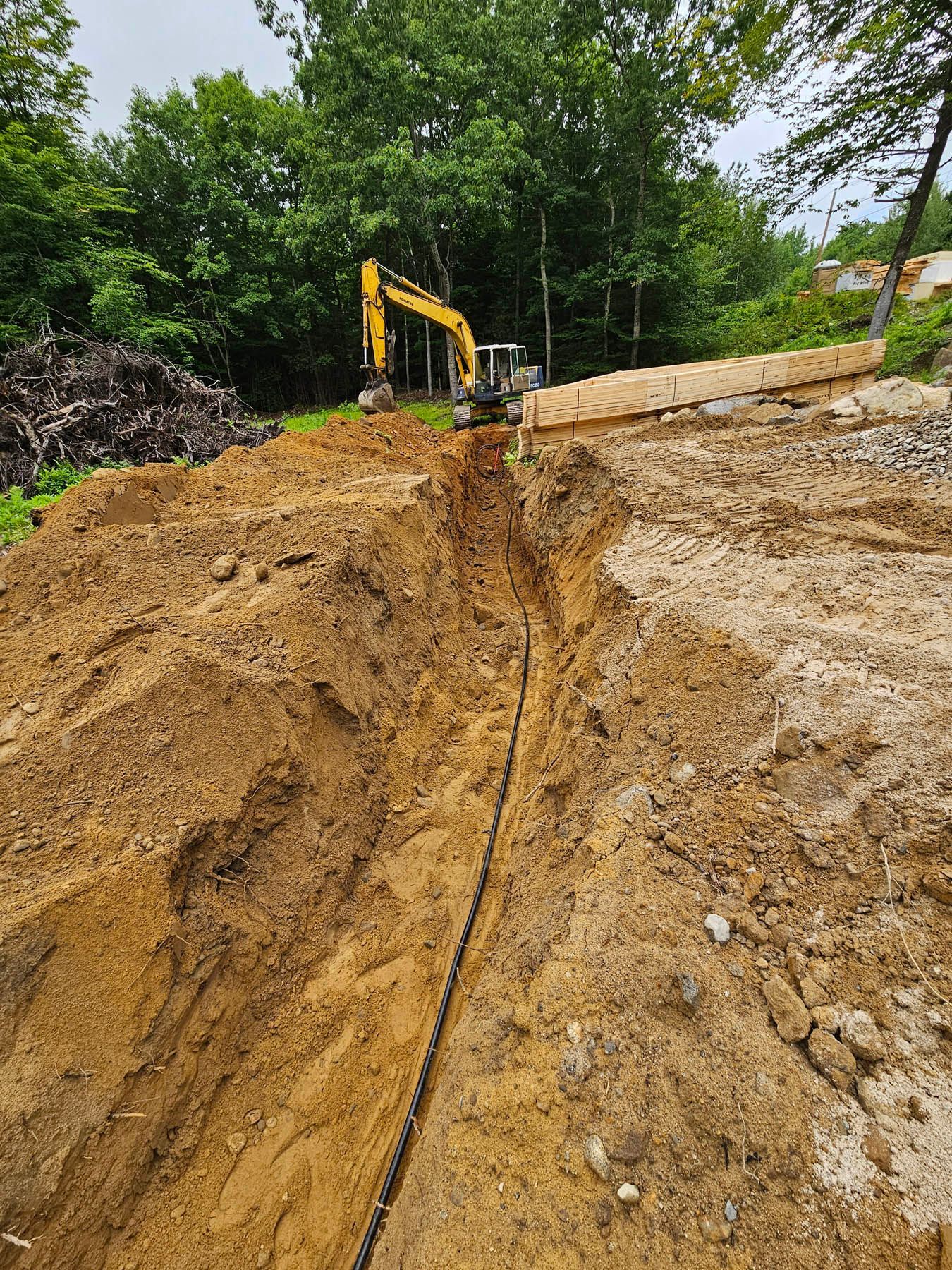 Excavator digging a trench in a hillside. A black cable runs along the trench. Green trees in the background.
