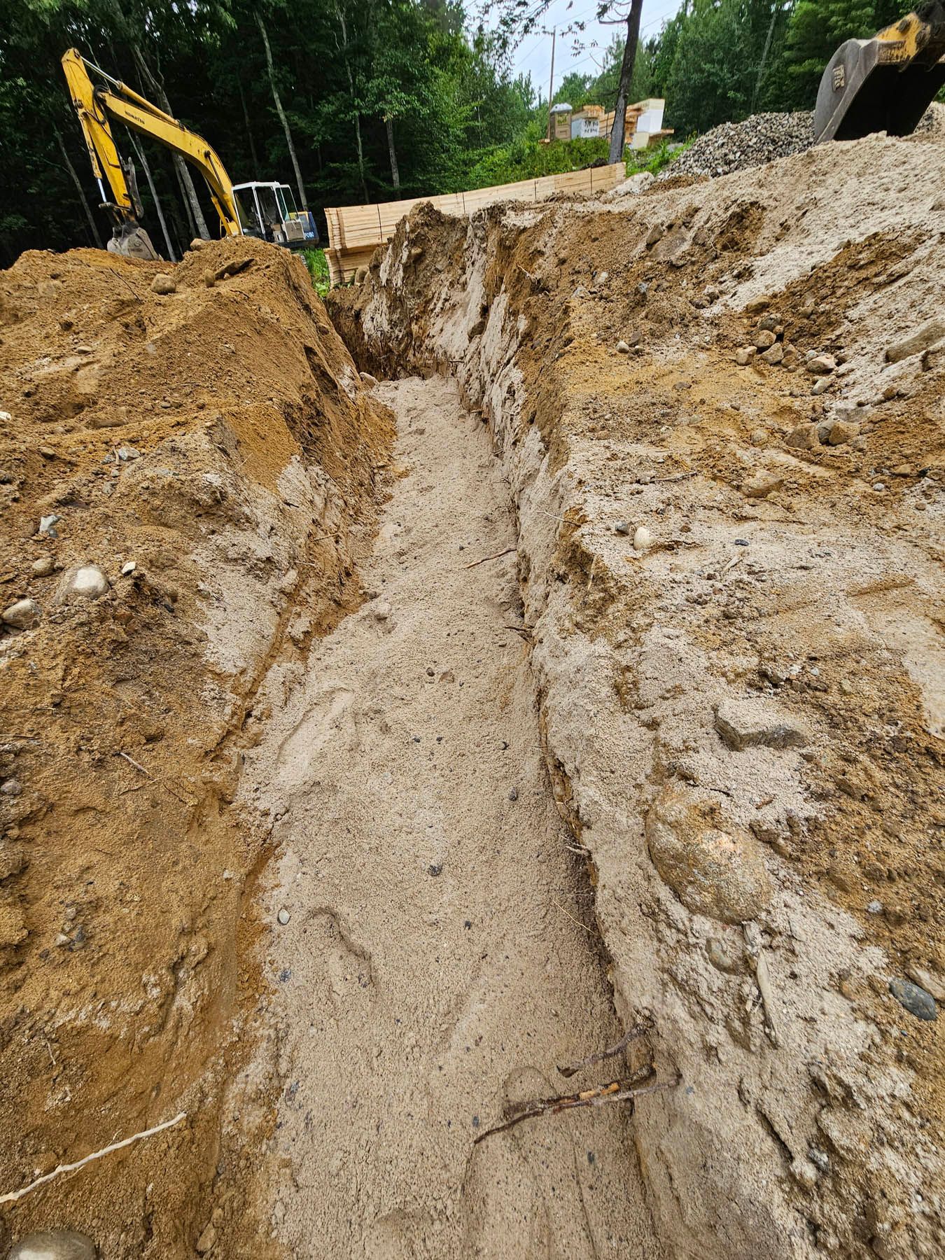Trench dug in dirt, filled with sand, construction site. Yellow excavator in background.