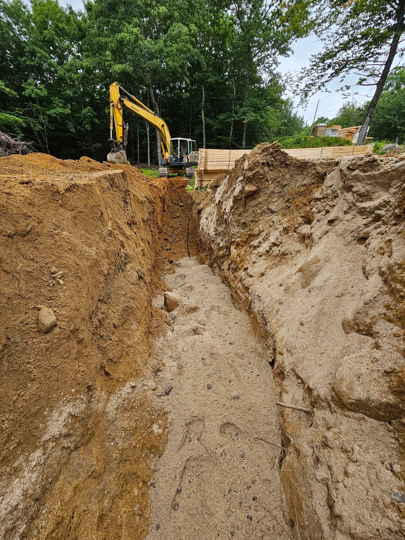 Dug trench with excavator in background, likely construction site, in a wooded area.