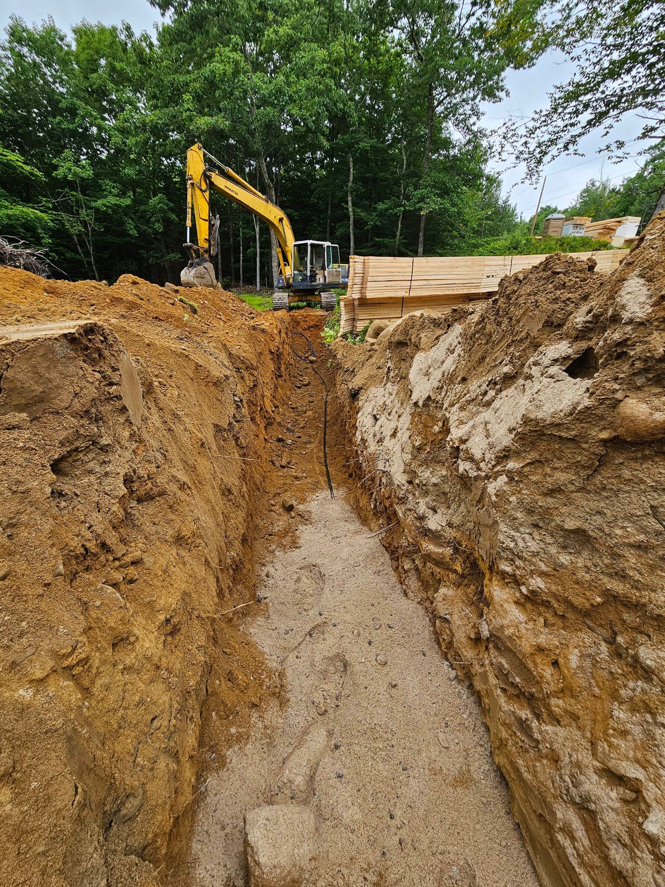 Ditch in earth with excavator in background, woods behind.