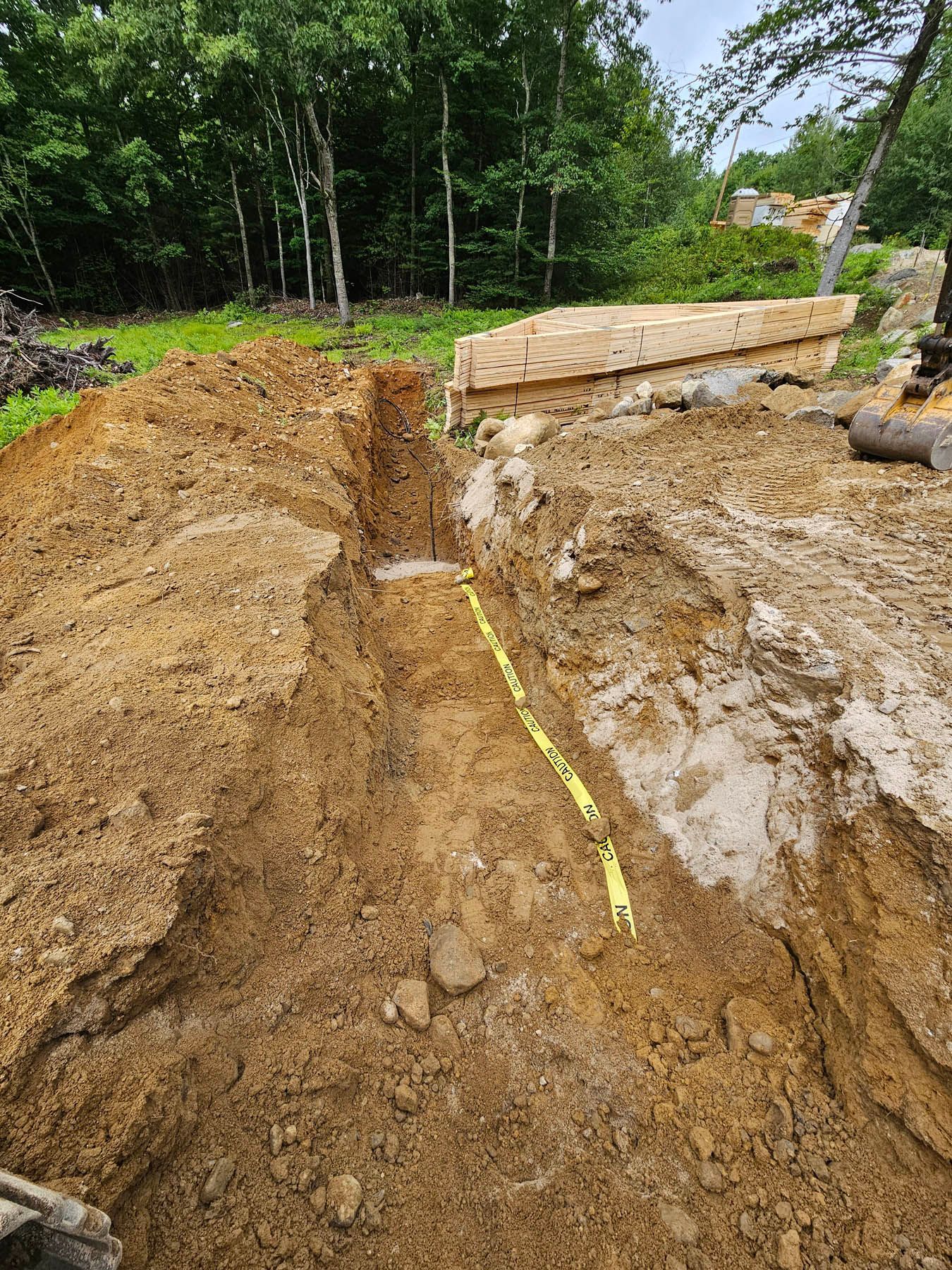 Trench dug in brown earth, with bright yellow gas line marking tape; trees and a wooden structure in the background.