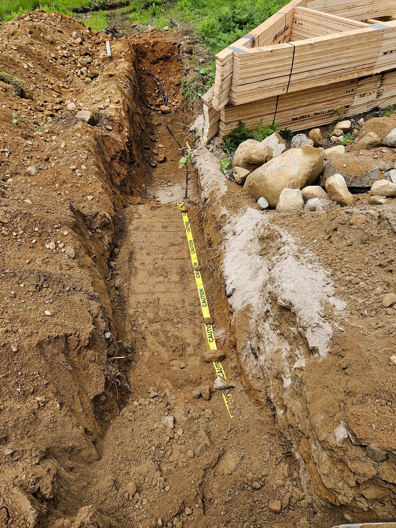 A narrow trench in dirt with a yellow measuring stick, near wood and rocks.