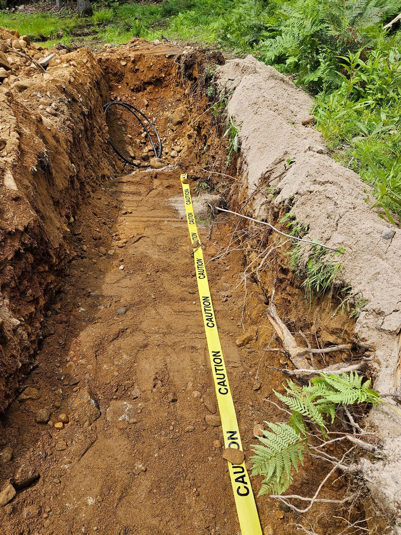 A dug trench with caution tape. Earth tones dominate, with green foliage visible on the right.