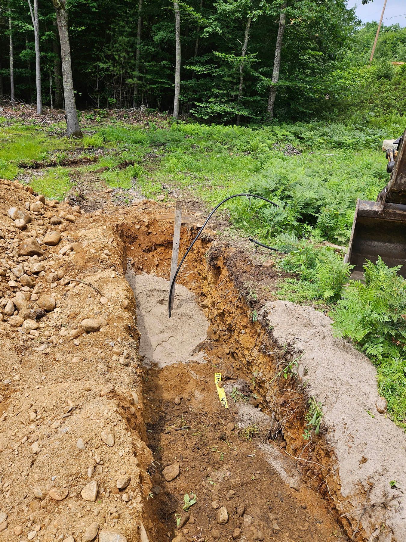 A trench dug in brown soil, with a black cable and yellow marker visible. Trees and vegetation in background.