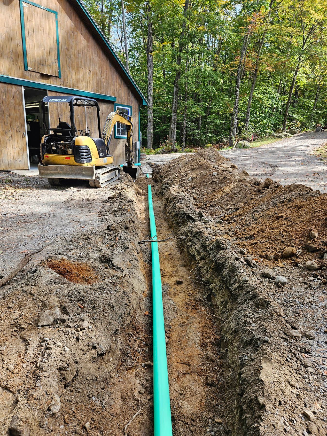 A trench dug for green pipe running from a barn; an excavator sits nearby.