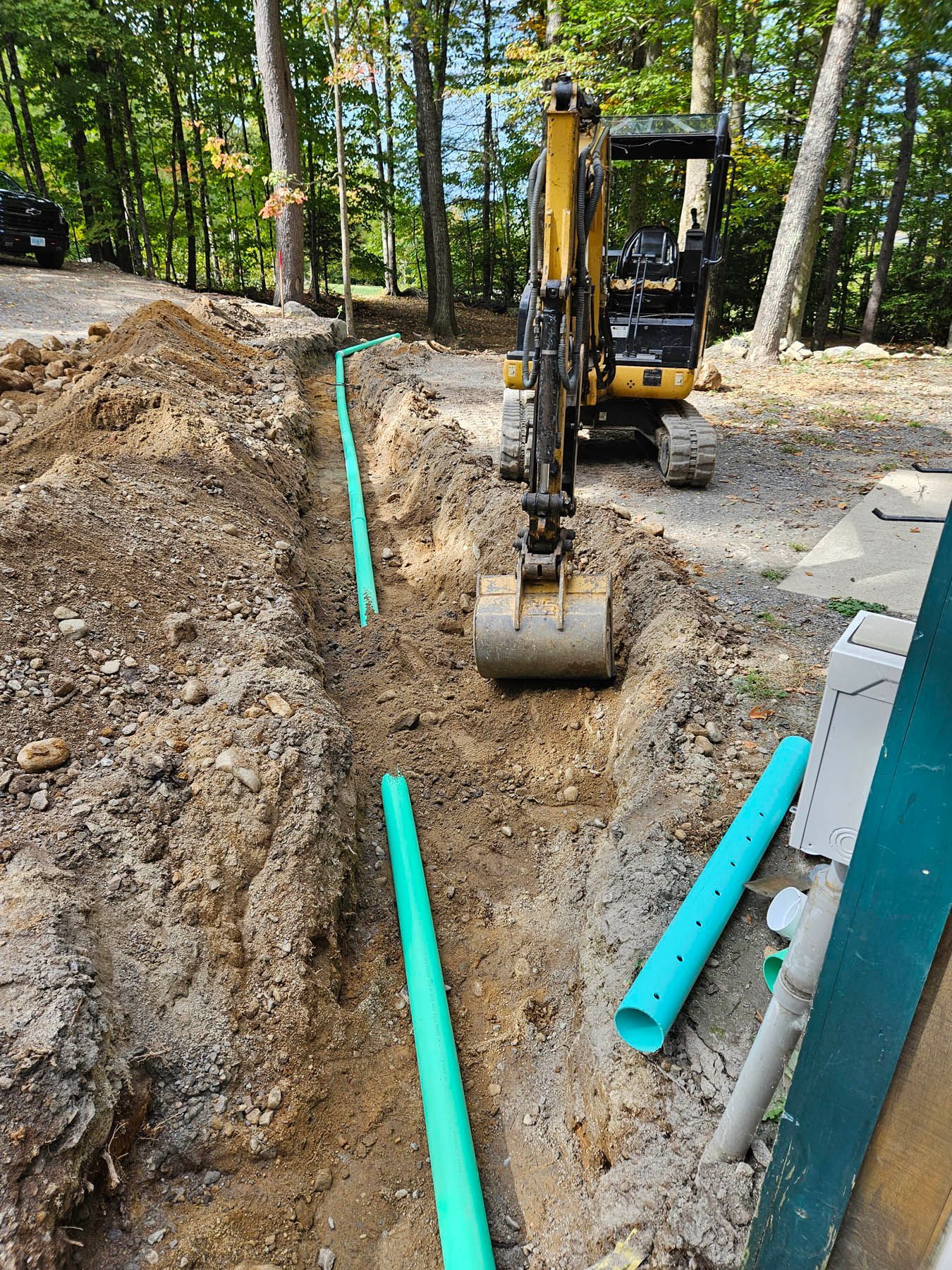 An excavator digging a trench for green pipes in a wooded area.