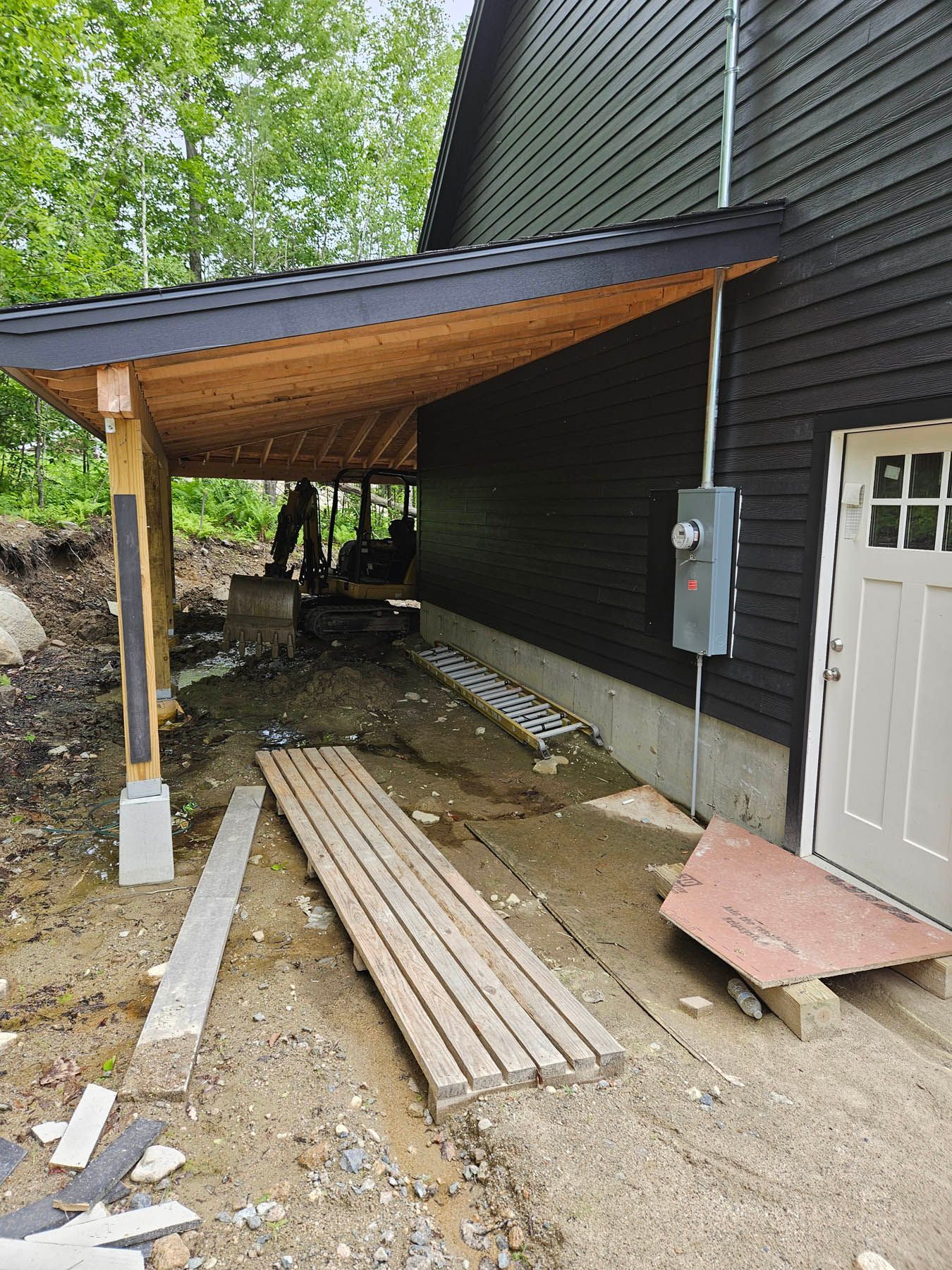 Construction of a covered walkway. Black building, wood under the roof. An excavator is visible in the background.