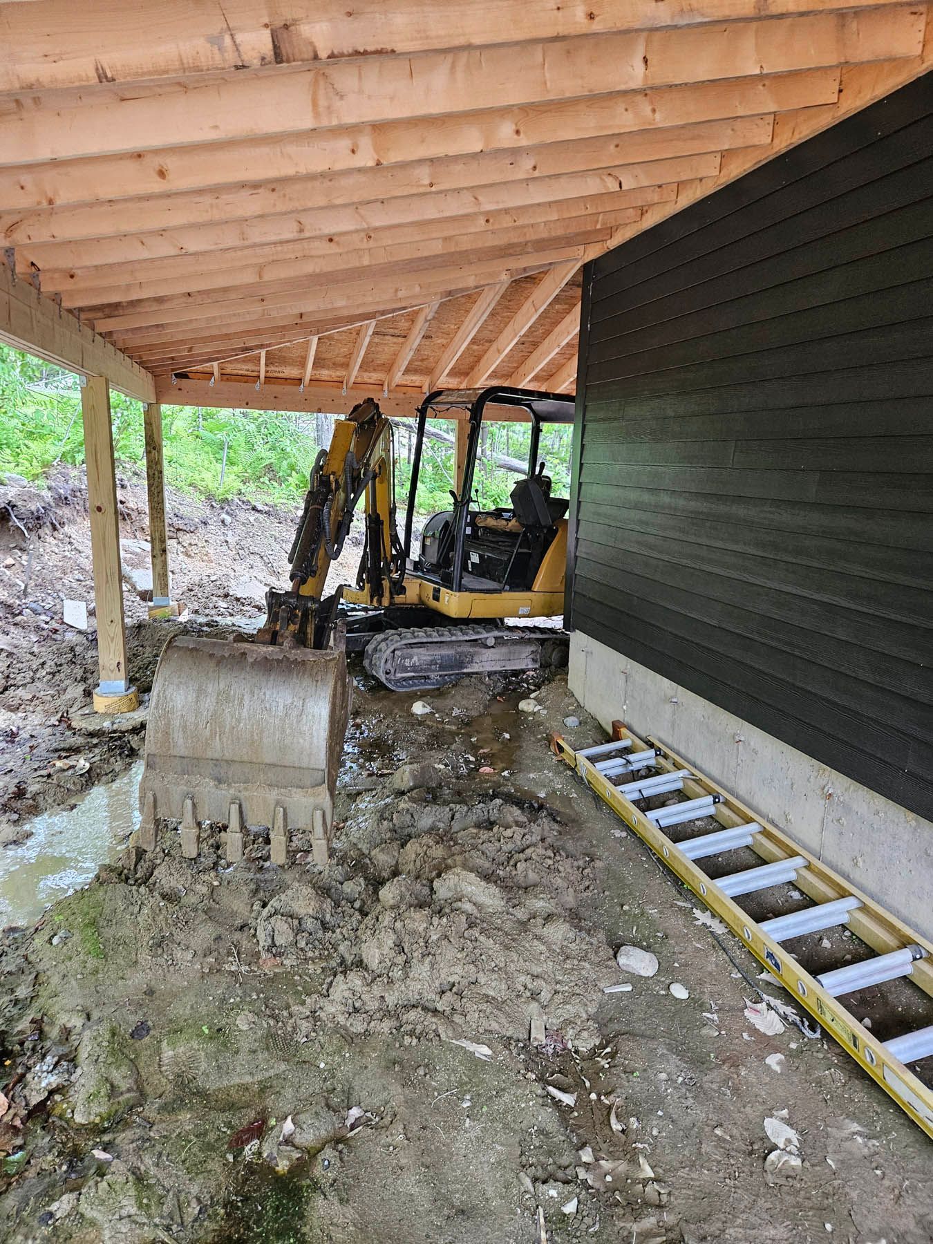 Mini excavator digging under a wooden canopy next to a dark wall; mud and a ladder present.