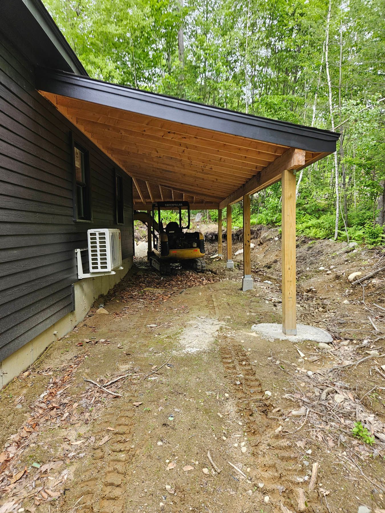 Lean-to roof attached to a dark-sided building, supported by wooden posts, covering an area with construction equipment.