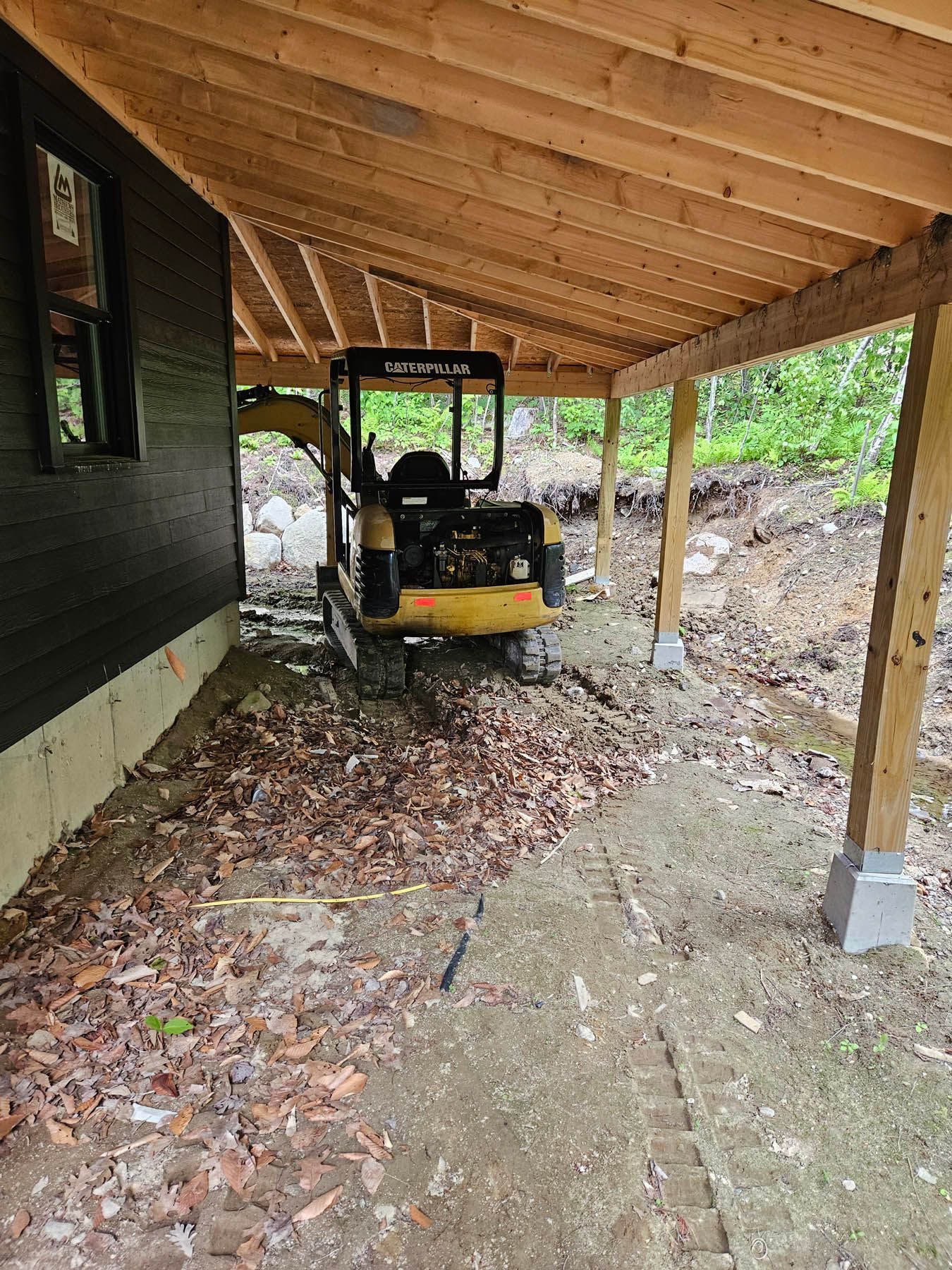 Yellow excavator parked under a wooden roofed structure near a dark-colored building.