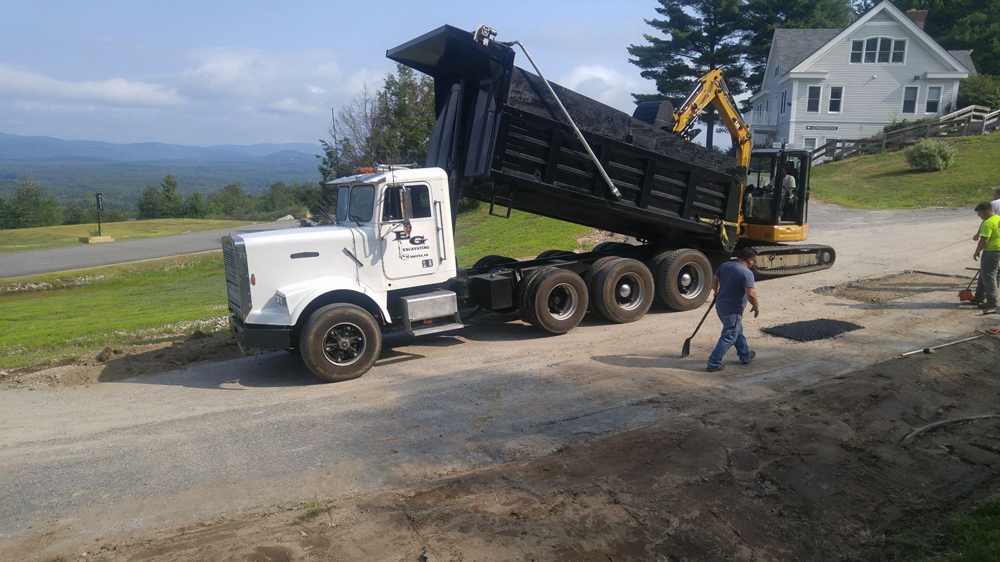 A white dump truck is being loaded by an excavator. Workers on a road in front of a white house.
