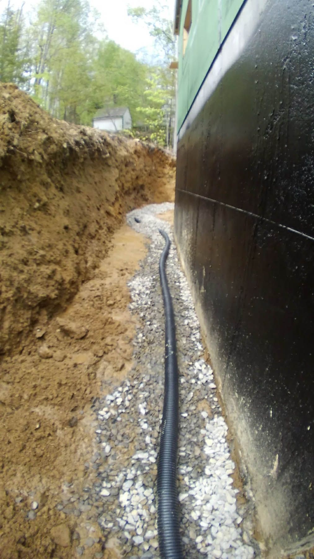 Trench with drainpipe along a black foundation wall, surrounded by gravel and dirt, outdoors.