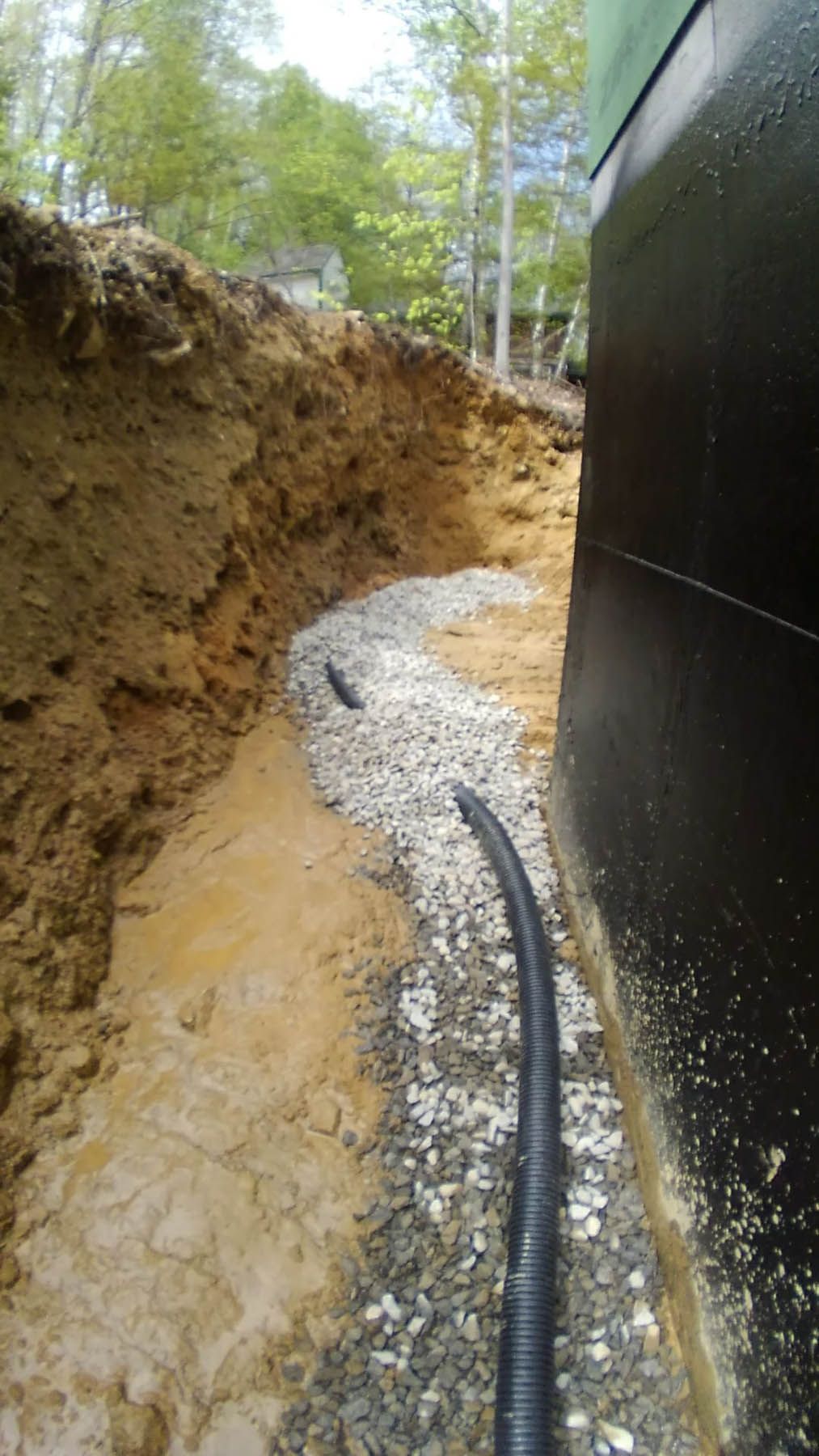 Gravel and black pipe line a trench along a black wall and dirt embankment. Sunlight filters through trees in the background.