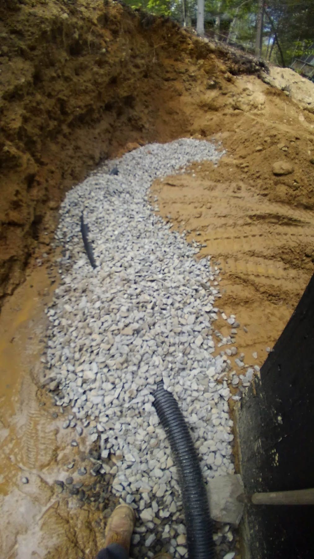 Gravel-filled trench with black drainage pipe along a hillside. Soil banks on either side.