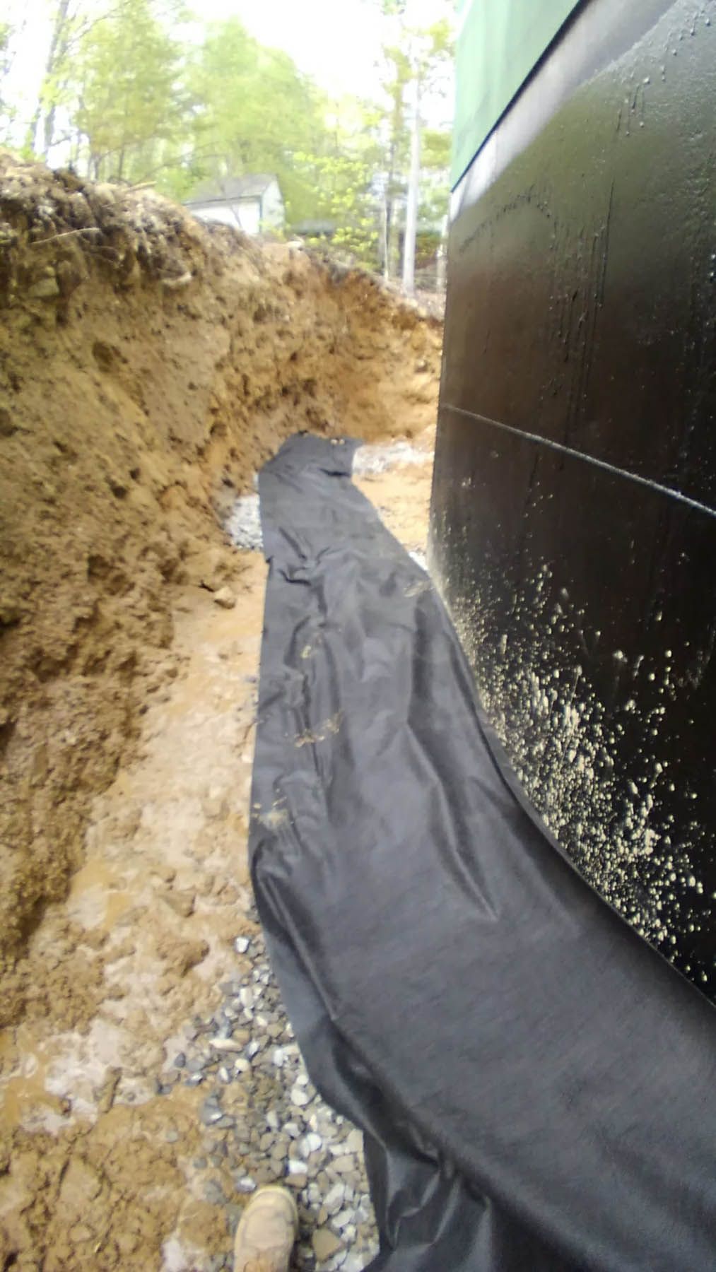 Black tar-coated foundation wall next to a dirt trench. Black fabric covers gravel drainage, outdoors.