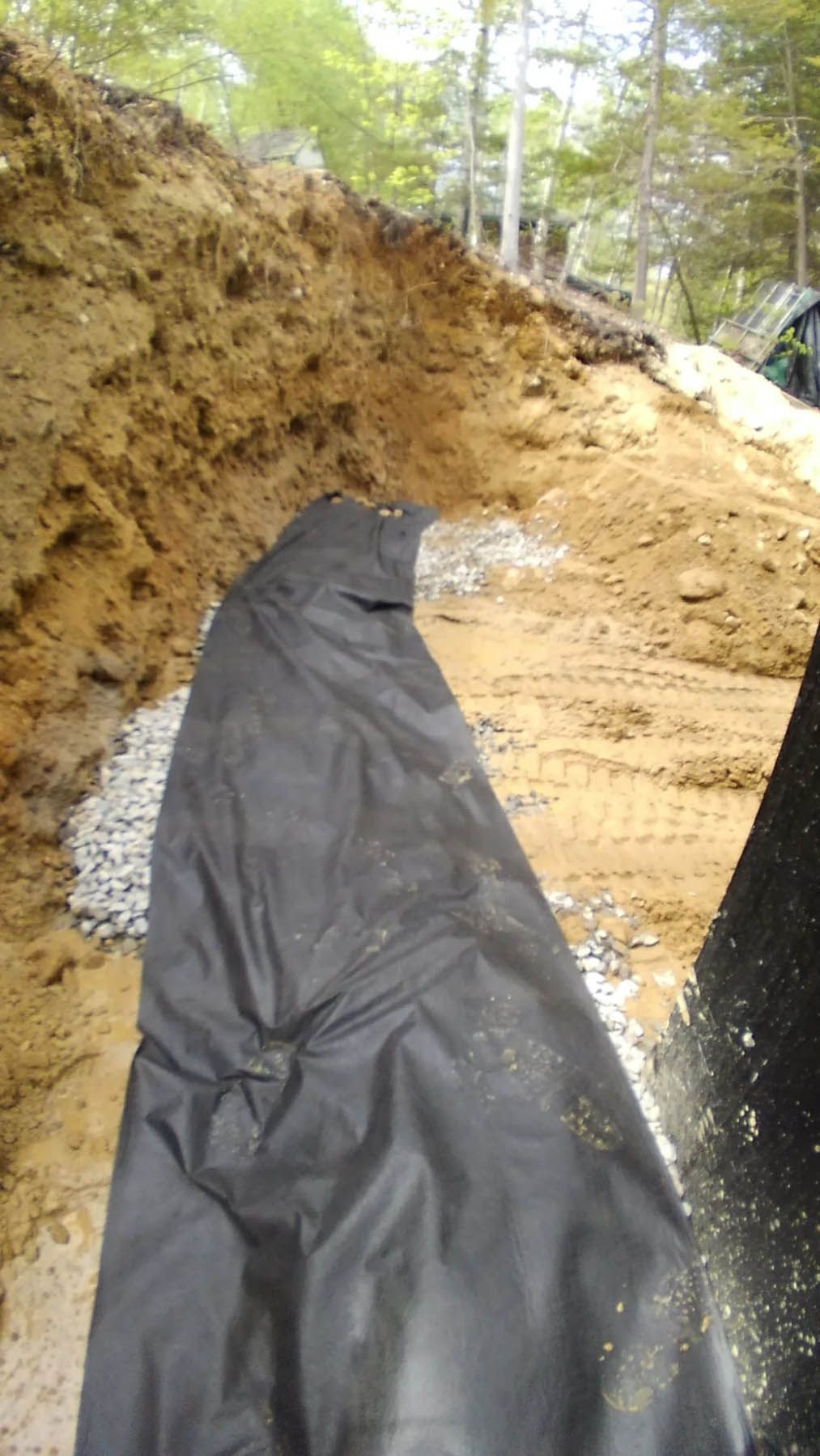 A dark fabric lining a trench with gravel, dirt embankment in background, trees in distance.