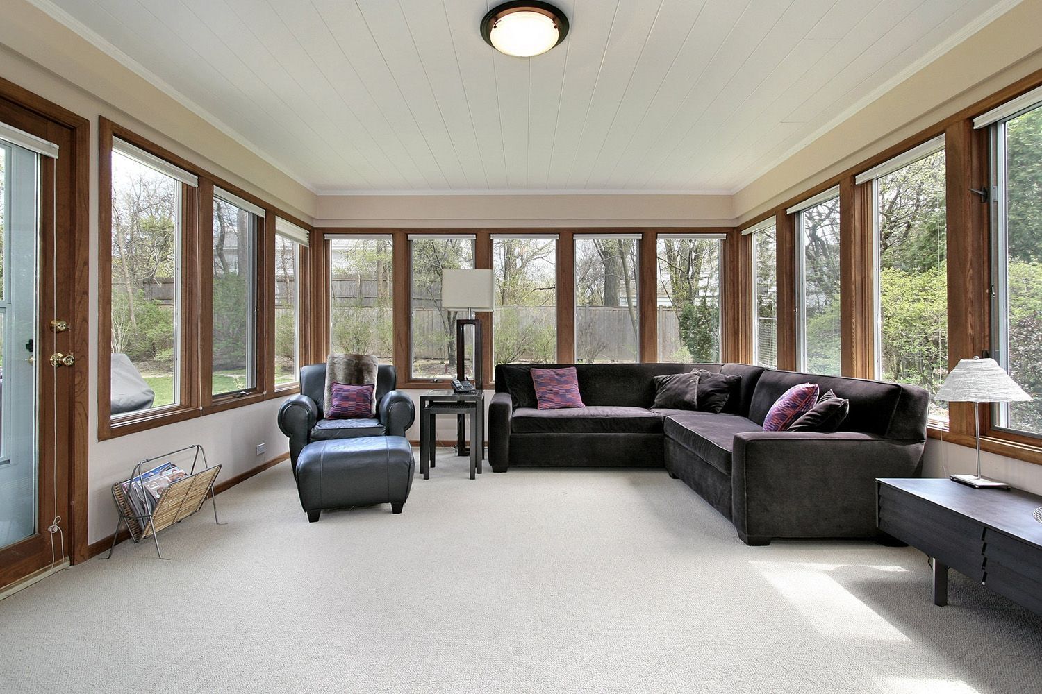 Sunroom with a dark L-shaped sofa and armchair, surrounded by windows. Beige carpet and a ceiling light.