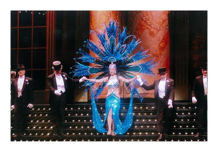 A woman in a blue feathered costume is surrounded by men in tuxedos