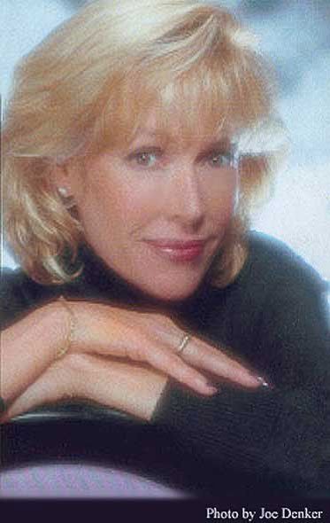 A woman with blonde hair is wearing a black turtleneck and a ring on her finger.