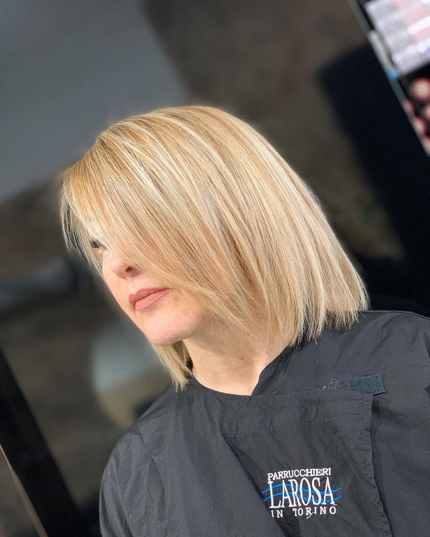 Donna con caschetto biondo e frangia, sguardo laterale. Indossa un top nero in un salone di bellezza.