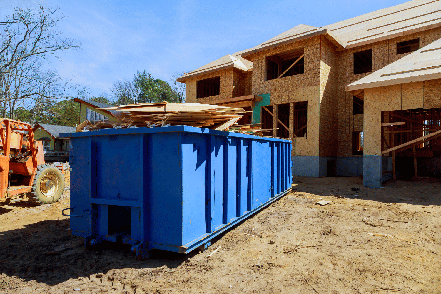 Blue construction dumpster filled with wood debris next to a wood-framed house under construction.