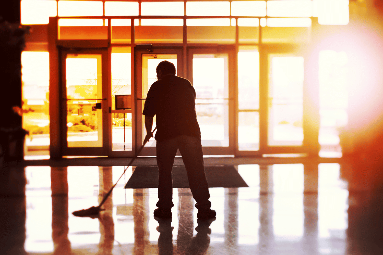 Person mopping a shiny floor in front of glass doors, backlit by bright sunlight.