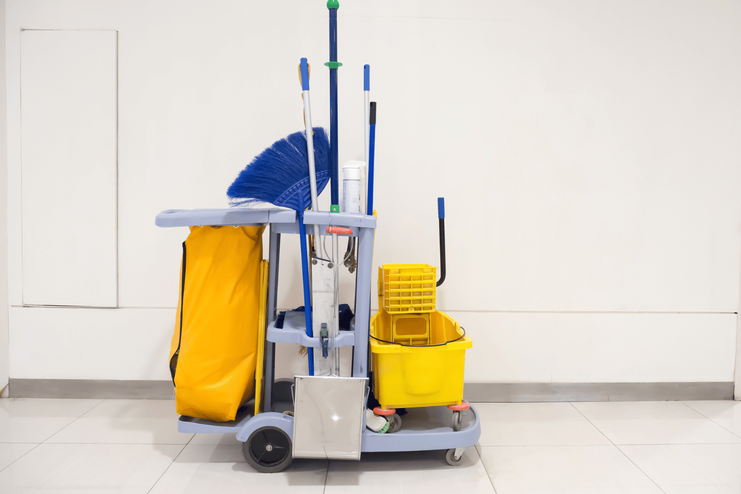 Cleaning cart with yellow bucket, blue broom, and cleaning supplies against a white wall.