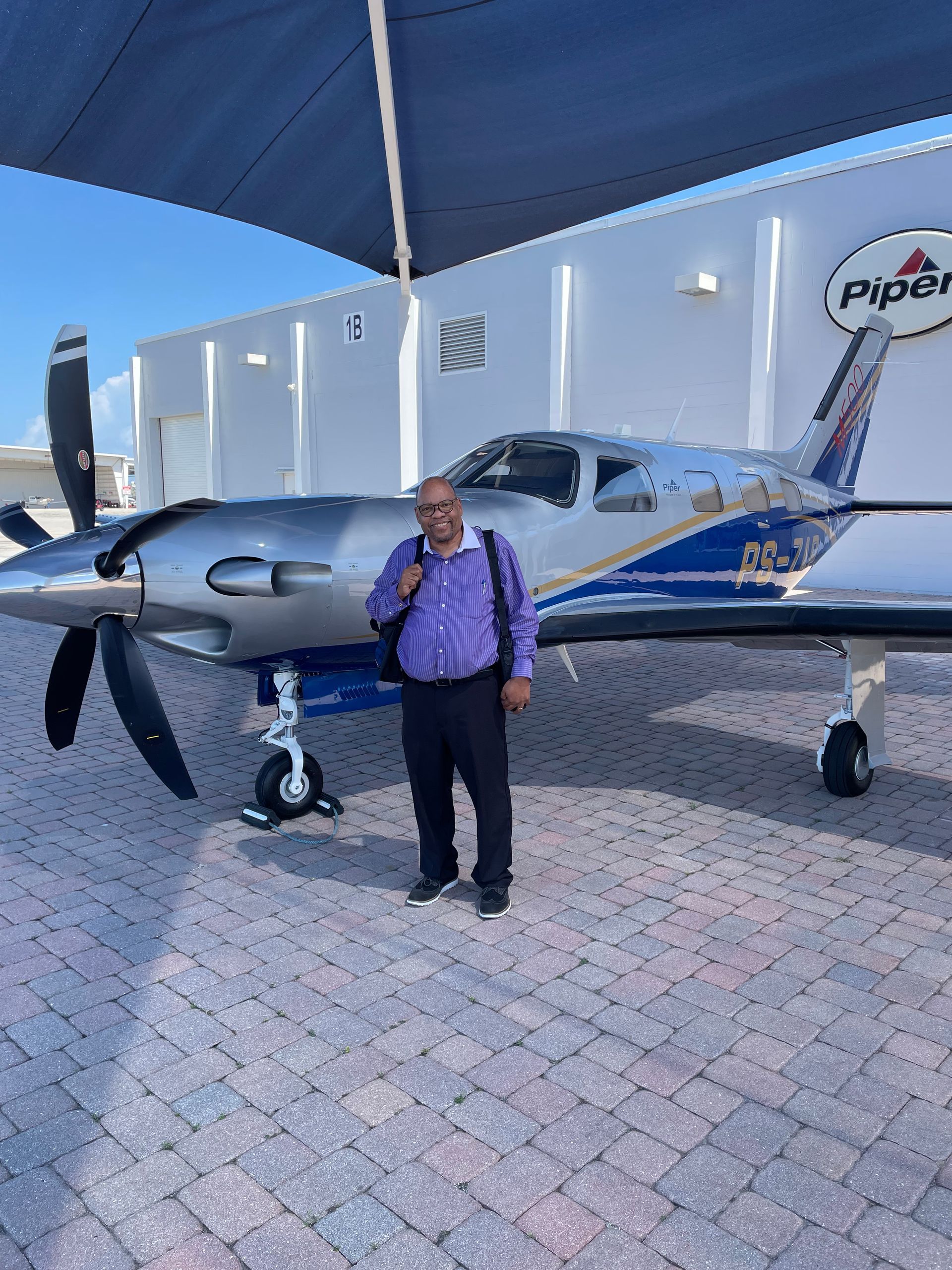 Man stands in front of a silver and blue Piper airplane; sunny day, paved area, white building.