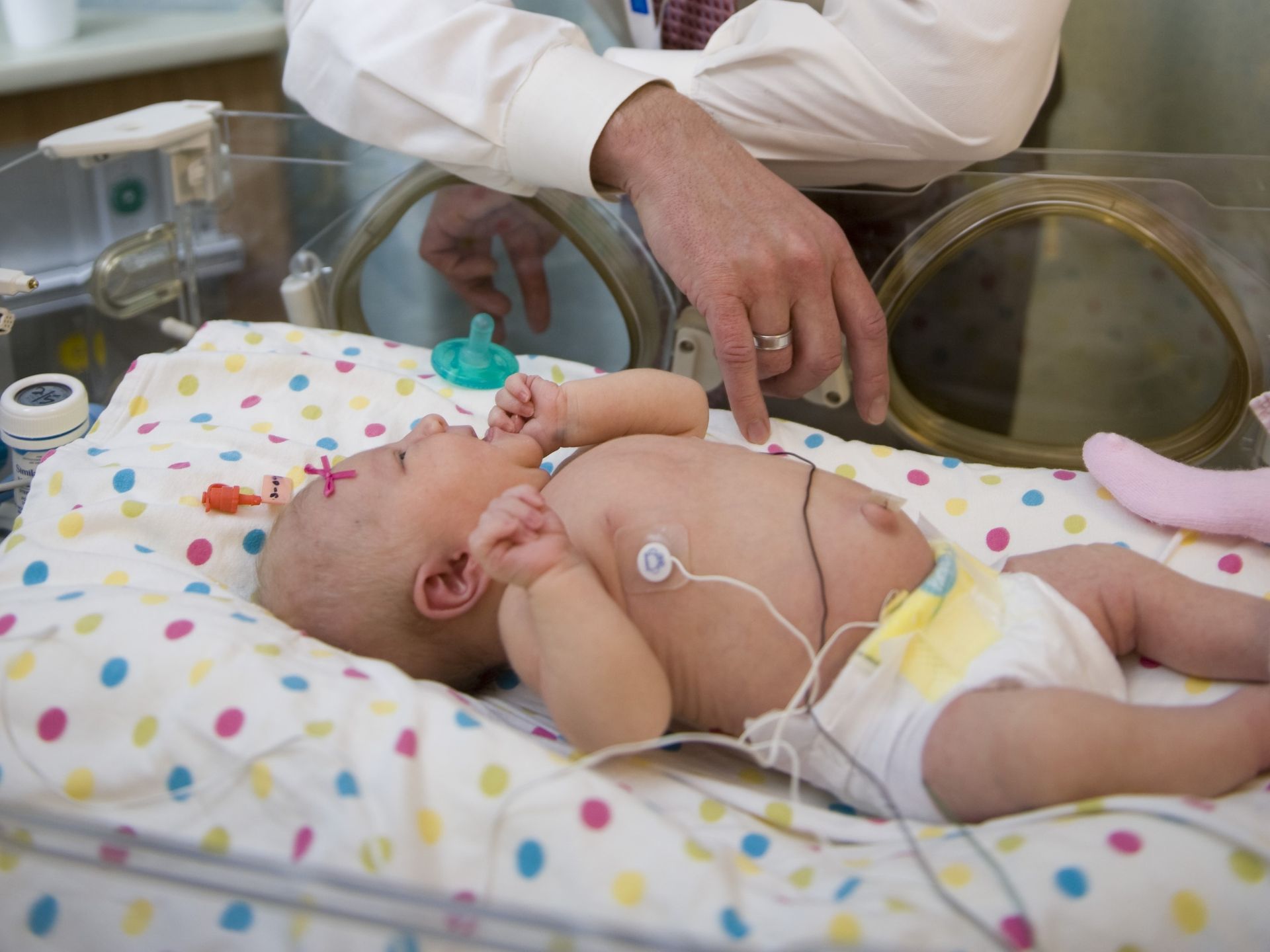 A baby in a hospital bed looks up at a doctor whose arms are visible above them.